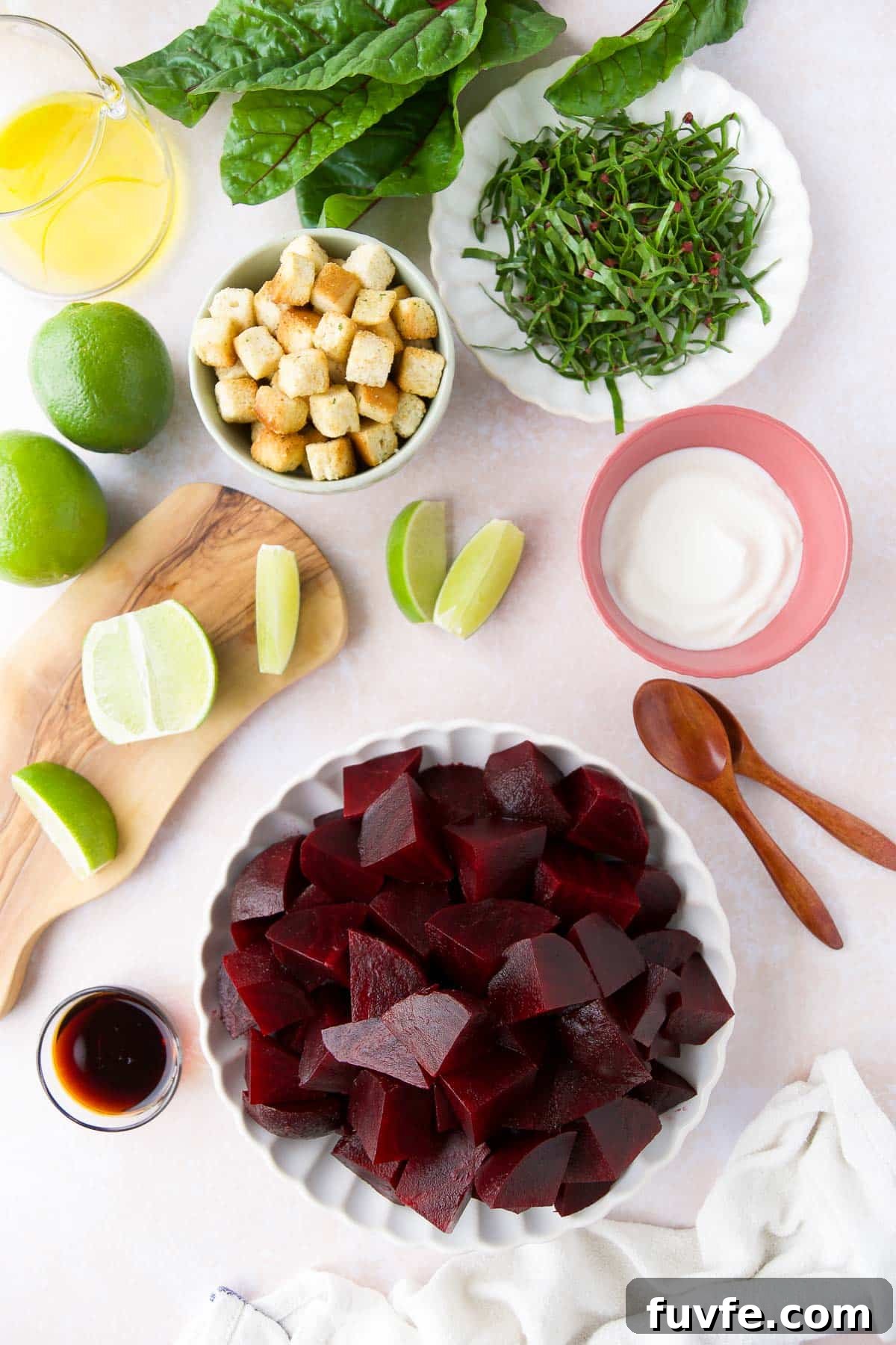 Ingredients for cold beet soup laid out: chopped cooked beets, Greek yogurt, croutons, sliced beet greens, lime wedges, soy sauce, and olive oil