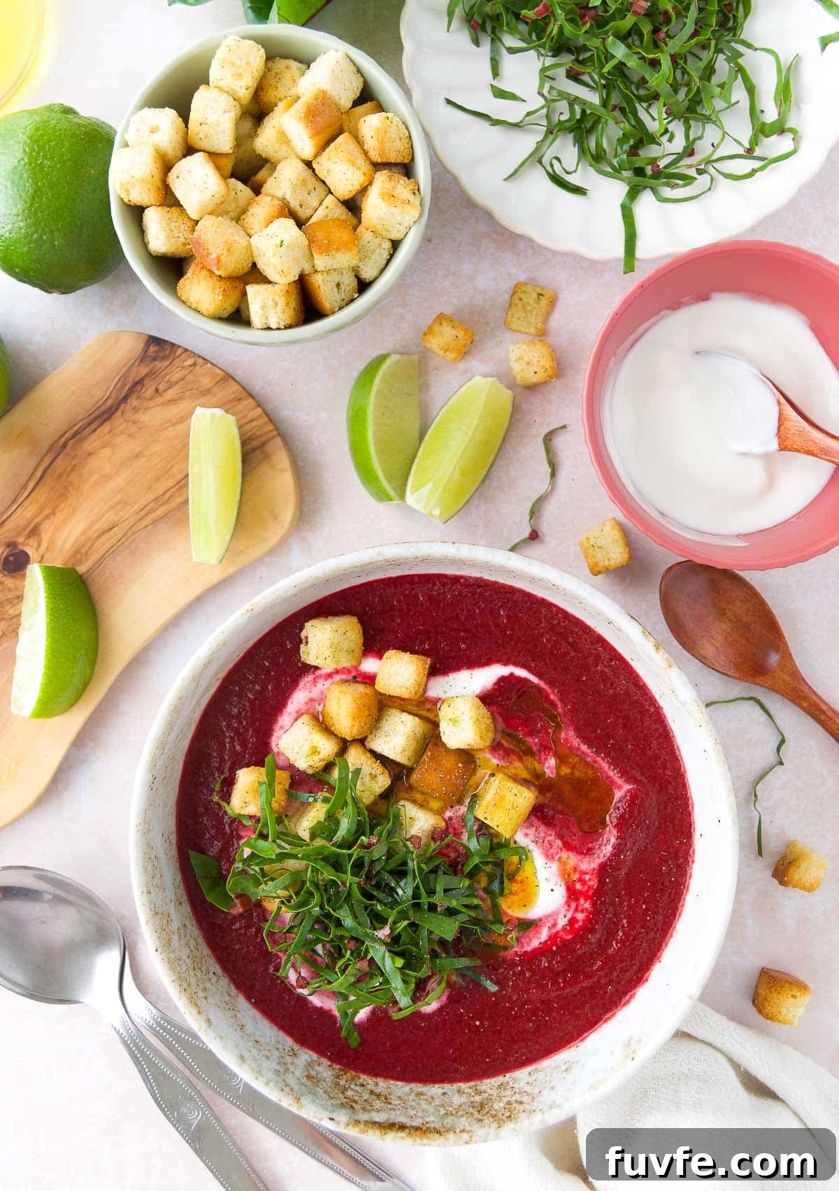 Cold beet soup in a white bowl topped with Greek yogurt swirl, croutons, and beet greens, with lime wedges and yogurt on the side