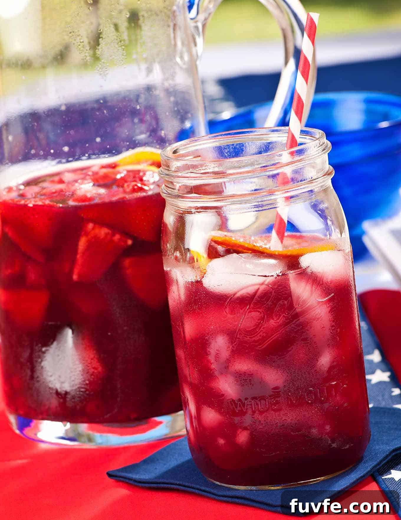 Refreshing Summer Berry Sangria Recipe for Backyard Parties 2 A mason jar filled with summer berry sangria with a red and white striped straw in it, a pitcher with more sangria with fruit in it in the background.