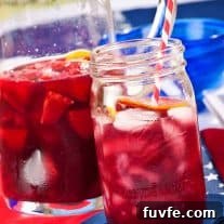 Refreshing Summer Berry Sangria Recipe for Backyard Parties 3 A mason jar filled with summer berry sangria with a red and white striped straw in it, a pitcher with more sangria with fruit in it in the background.