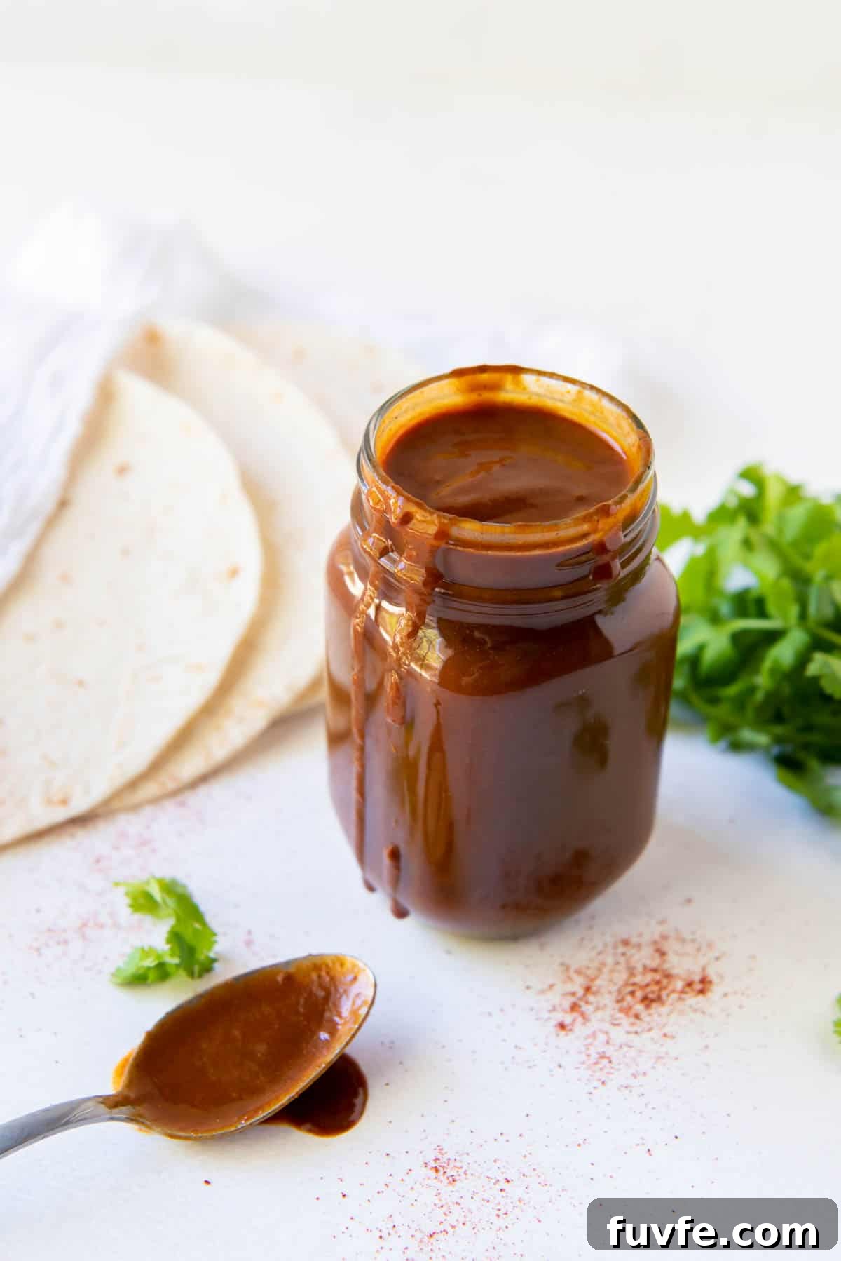 Homemade enchilada sauce in a glass jar with tortillas in the background and a spoon of sauce in front.