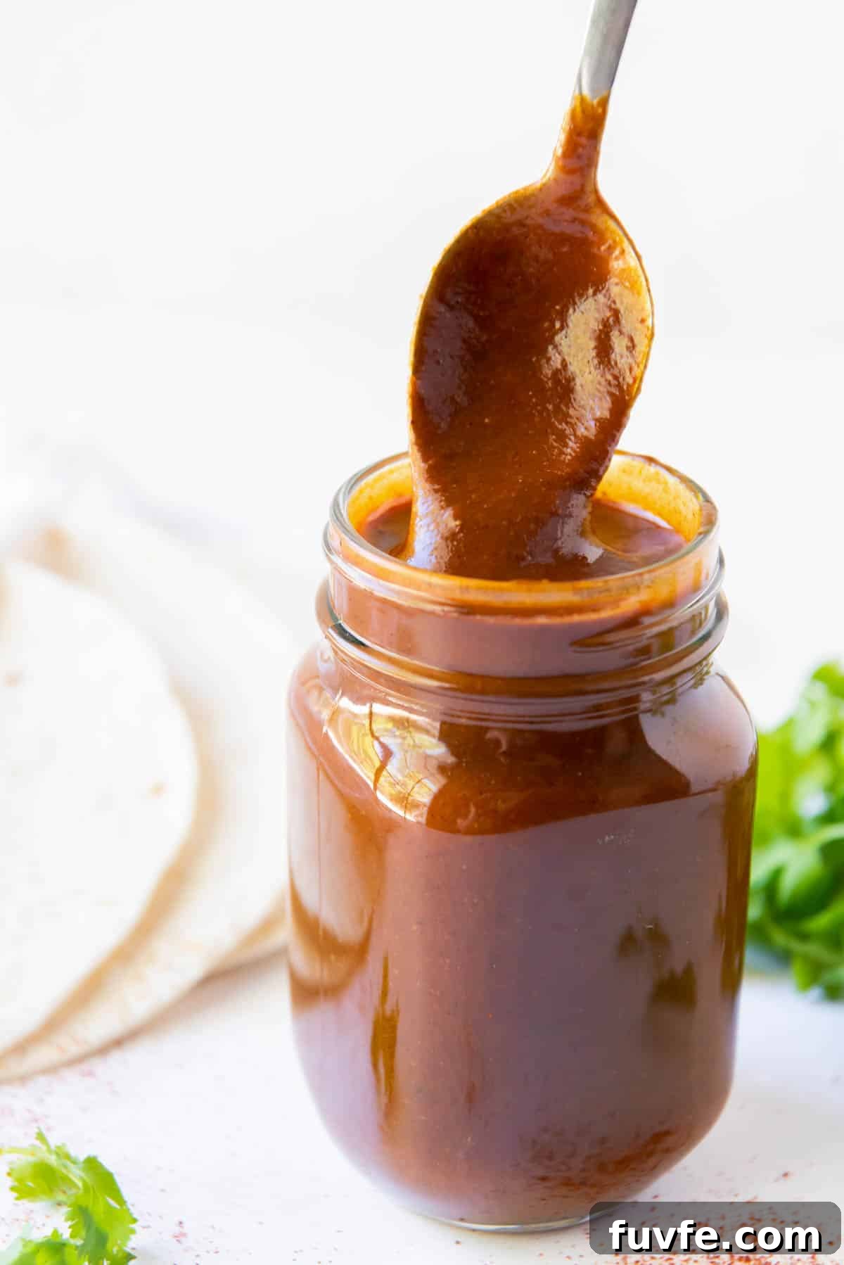 A glass jar of homemade enchilada sauce with a spoon stirring the sauce.