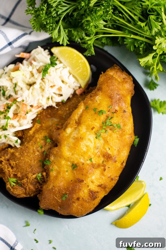 Two pieces of beer battered catfish on a plate with creamy coleslaw and a lemon wedge with fresh parsley and lemon wedges in the background around it.