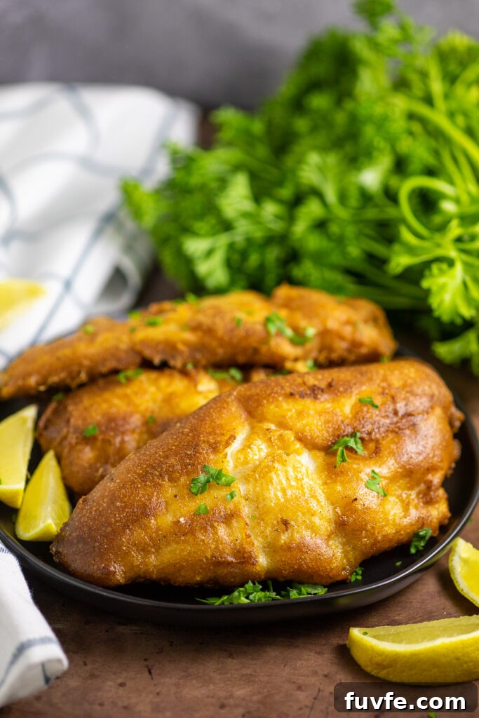 A black matte plate stacked with deep fried beer battered catfish topped with a sprinkle of fresh parsley with a clean linen and bunch of fresh parsley behind and lemon wedges in front.