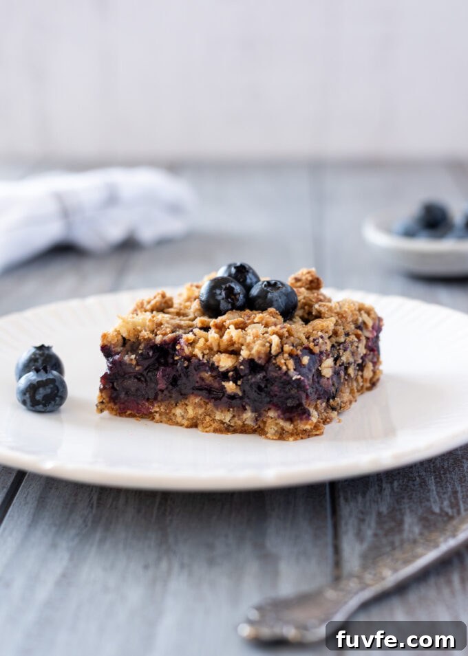 Blueberry Crumble Bars Recipe with Oat Crust 11 Blueberry crumble bar on a plate with fresh blueberries.