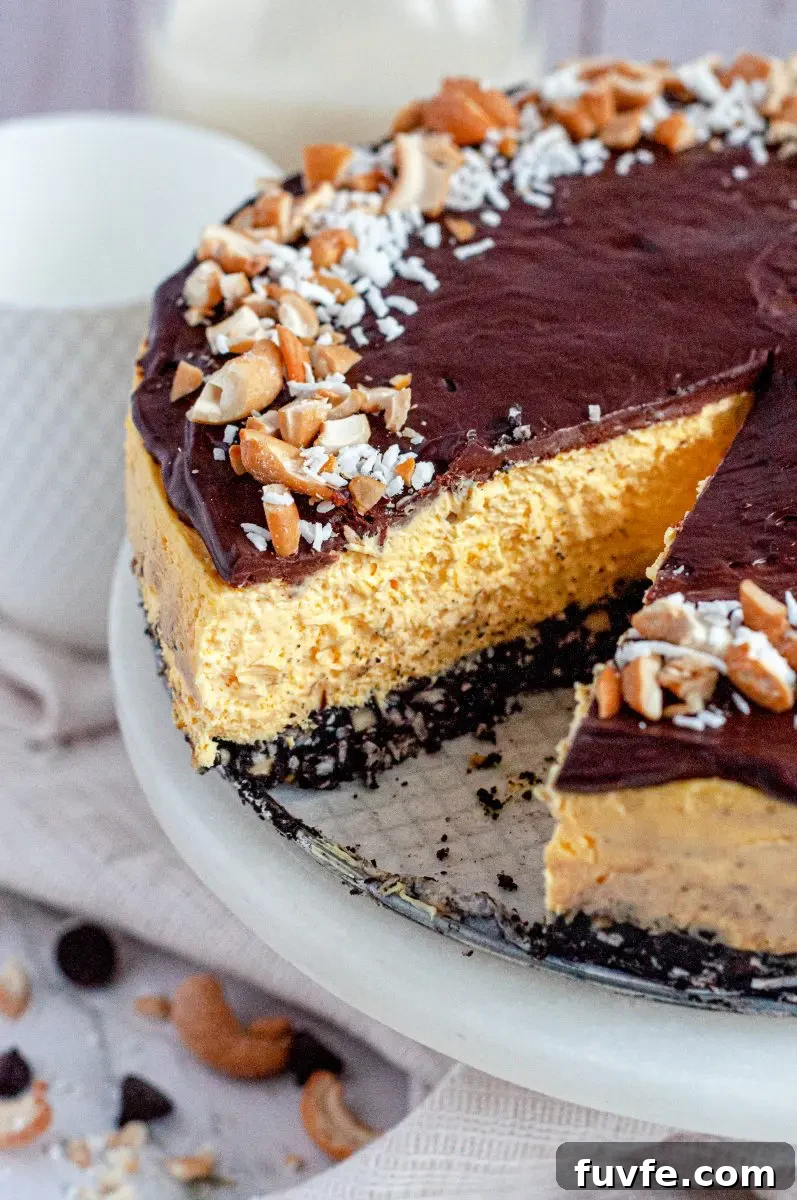 A no-bake nanaimo bar cheesecake. it has three layers. The bottom layer is a chocolate crust, the middle layer is a yellow custard cheesecake, and the top is a thin layer of dark chocolate topped with white shredded coconut and yellow salted cashews. There is a slice cut out of the cheesecake.