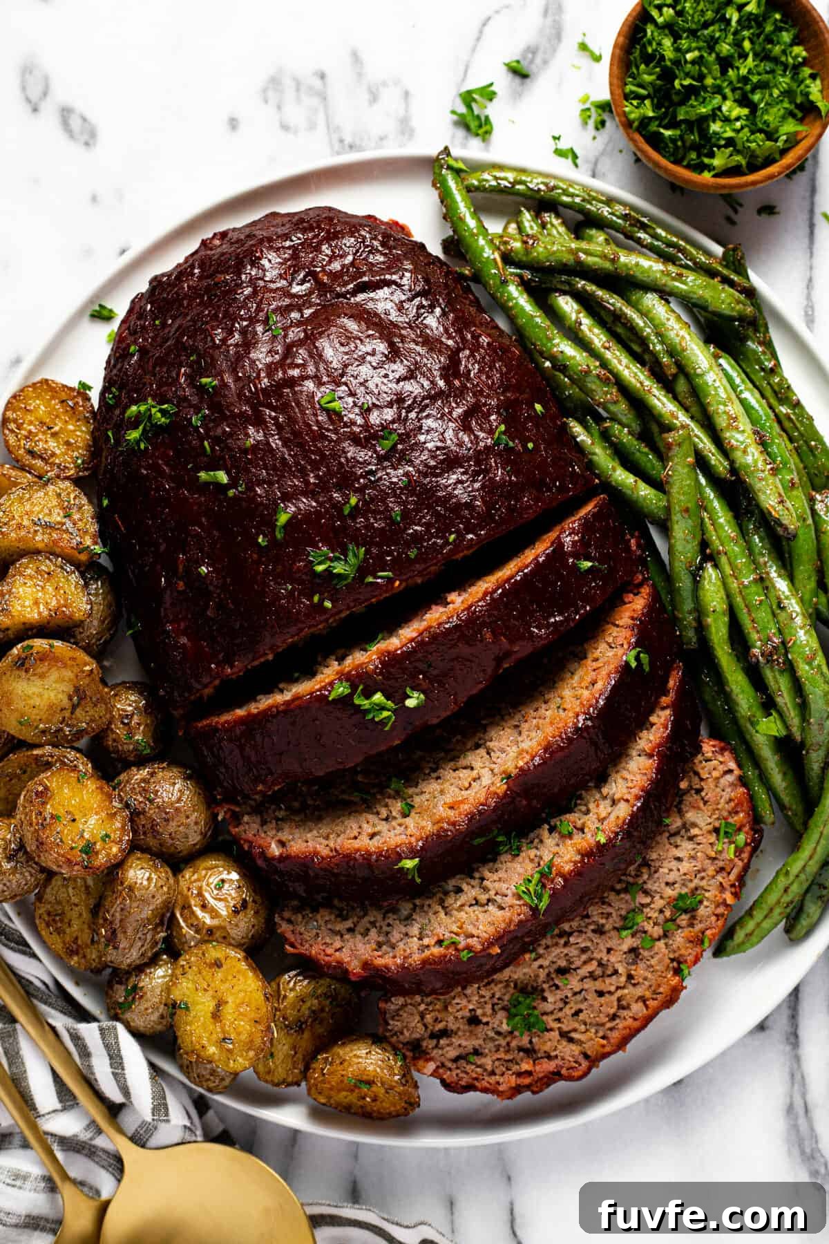 Sliced meatloaf on a white plate with roasted potatoes and green beans.