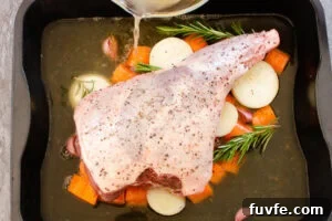 A leg of lamb in a roasting tin on chopped carrots and onions with stock being poured into the roasting tray