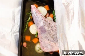 A leg of lamb in a roasting tin on chopped carrots and onions with stock being covered with aluminium foil