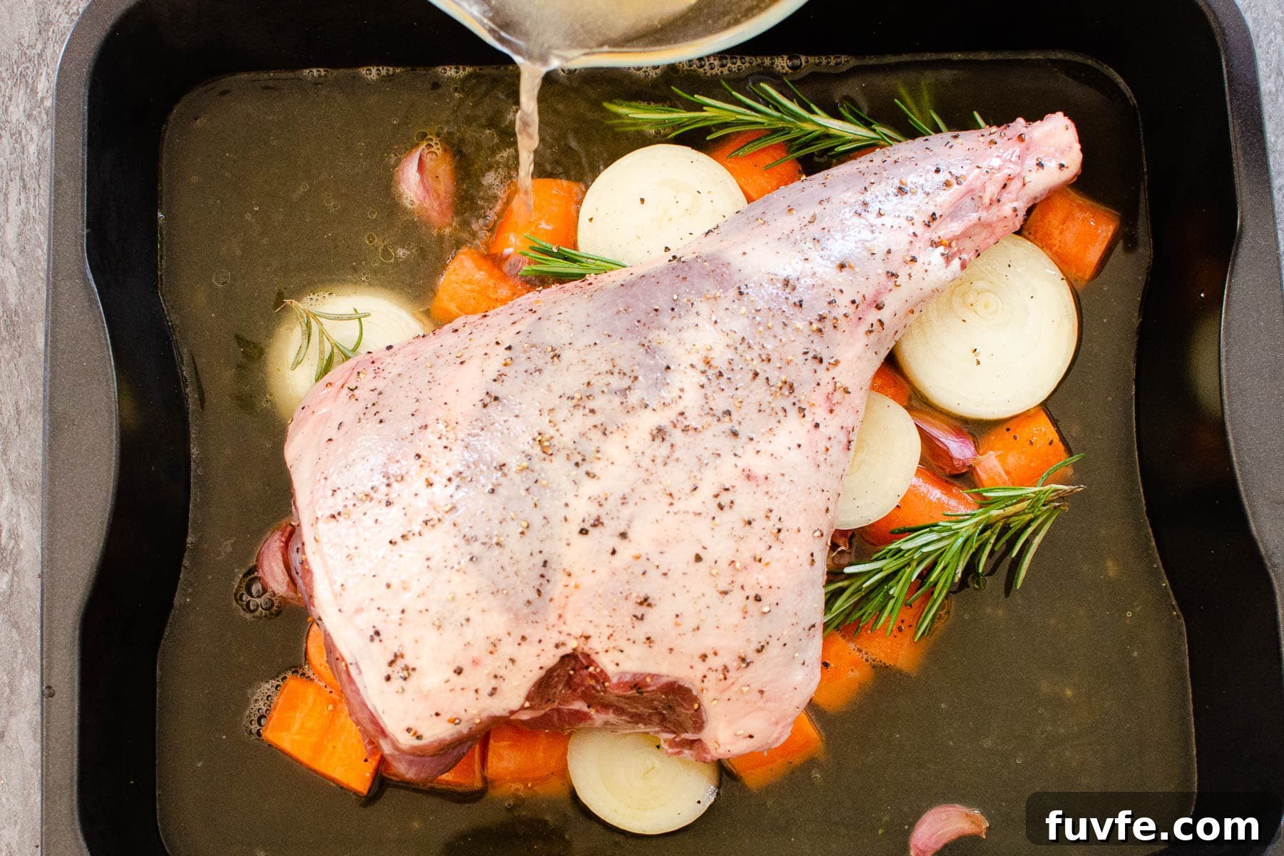 A leg of lamb in a roasting tin on chopped carrots and onions with stock being poured into the roasting tray