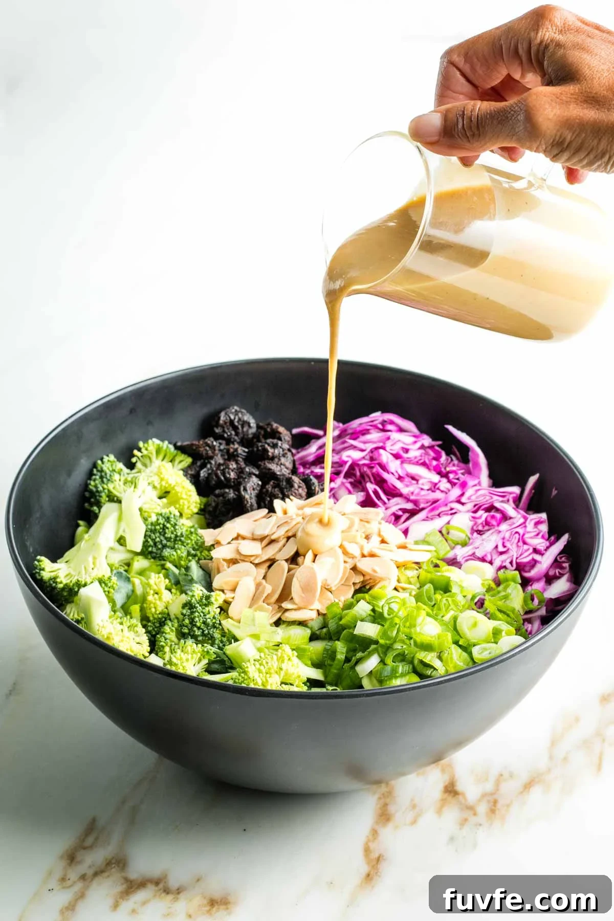 Pouring miso dressing over raw broccoli, cabbage, scallions plus almonds and dried cherries in a bowl.