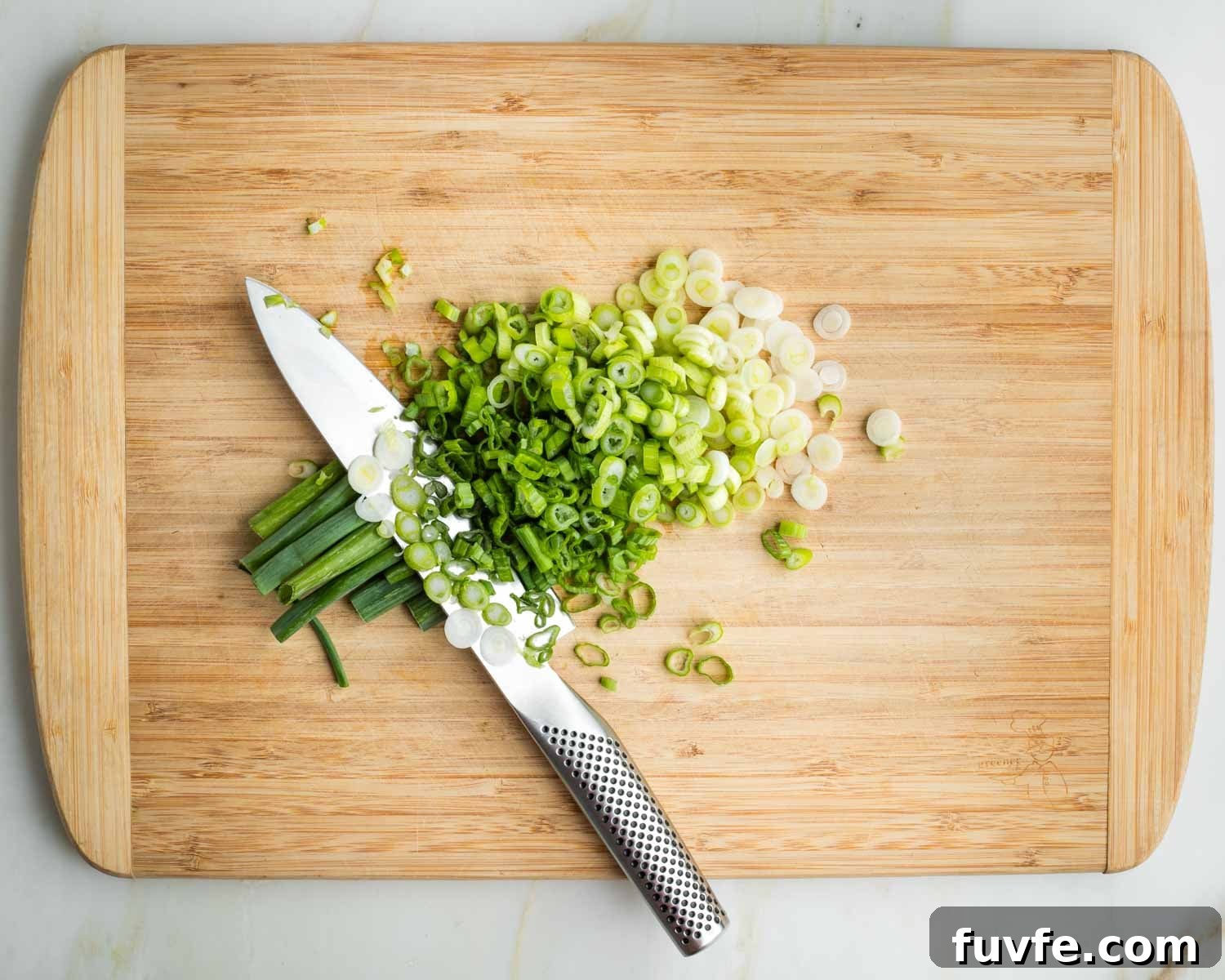 Thinly slicing white and green parts of green onions, on a wooden board.