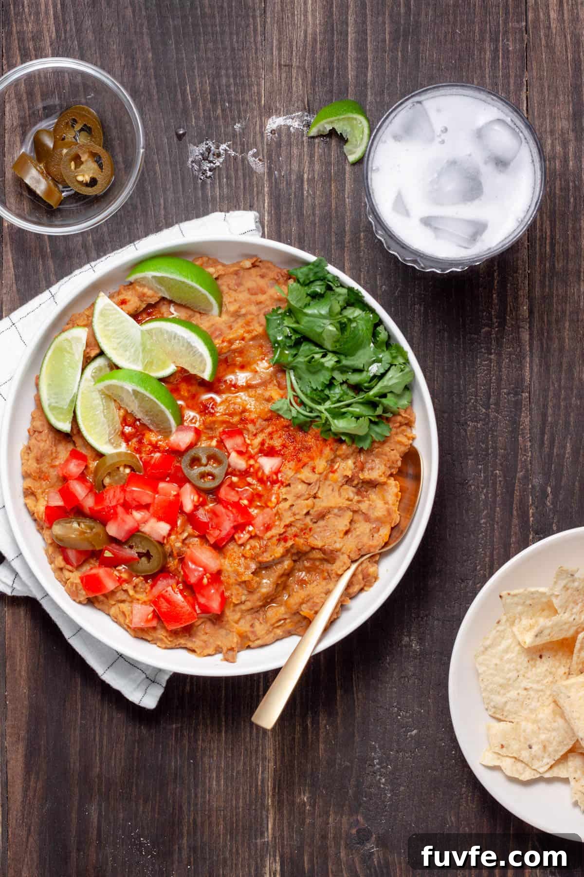 Vegan refried beans in a serving dish topped with lime wedges, cilantro leaves, chopped tomatoes and pickled jalapenos. 