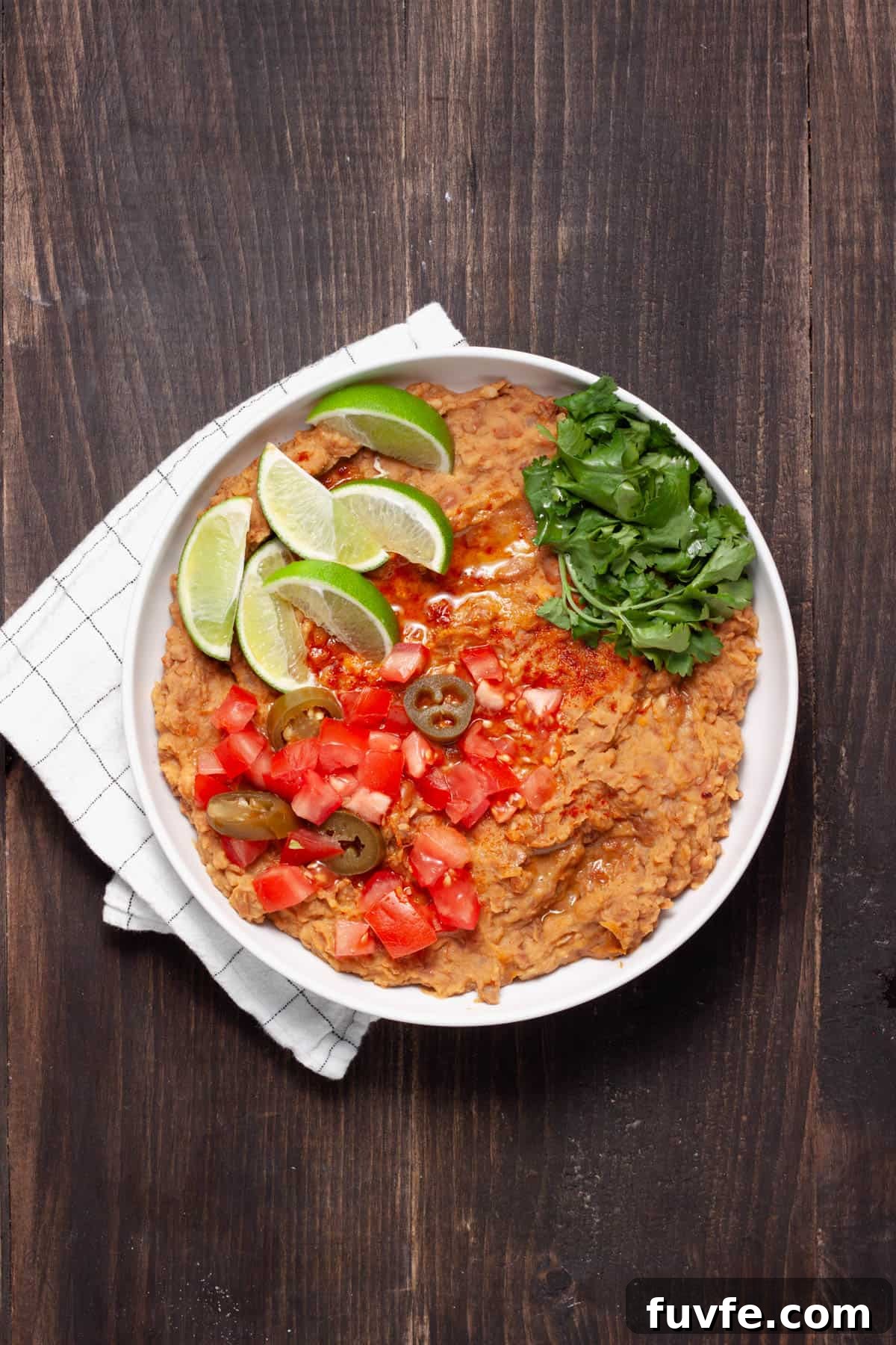 A bowl of vegan refried beans topped with cilantro, tomatoes, lime wedges and pickled jalapenos.