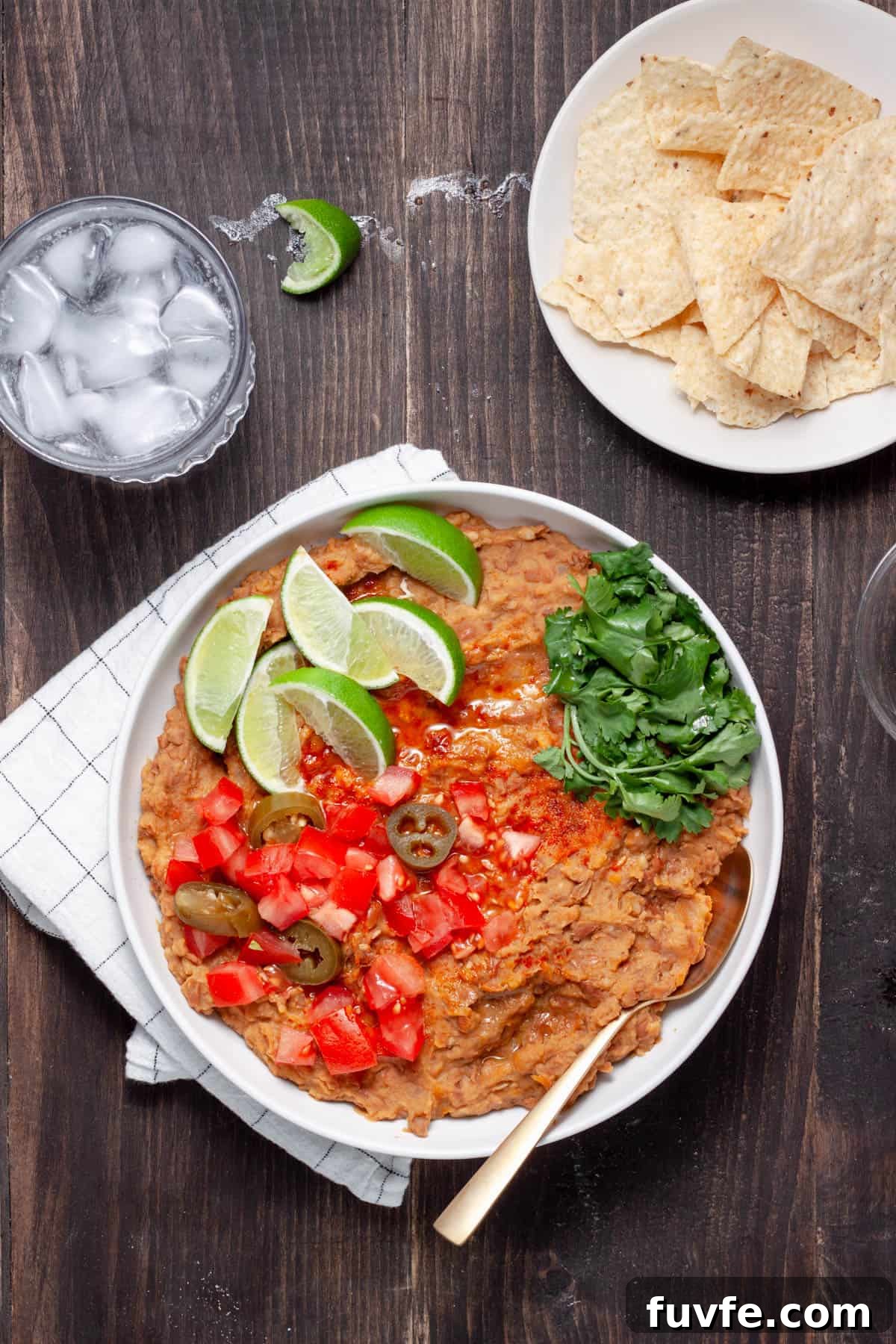 Vegan refried beans in a serving dish topped with lime wedges, cilantro leaves, chopped tomatoes and pickled jalapenos.