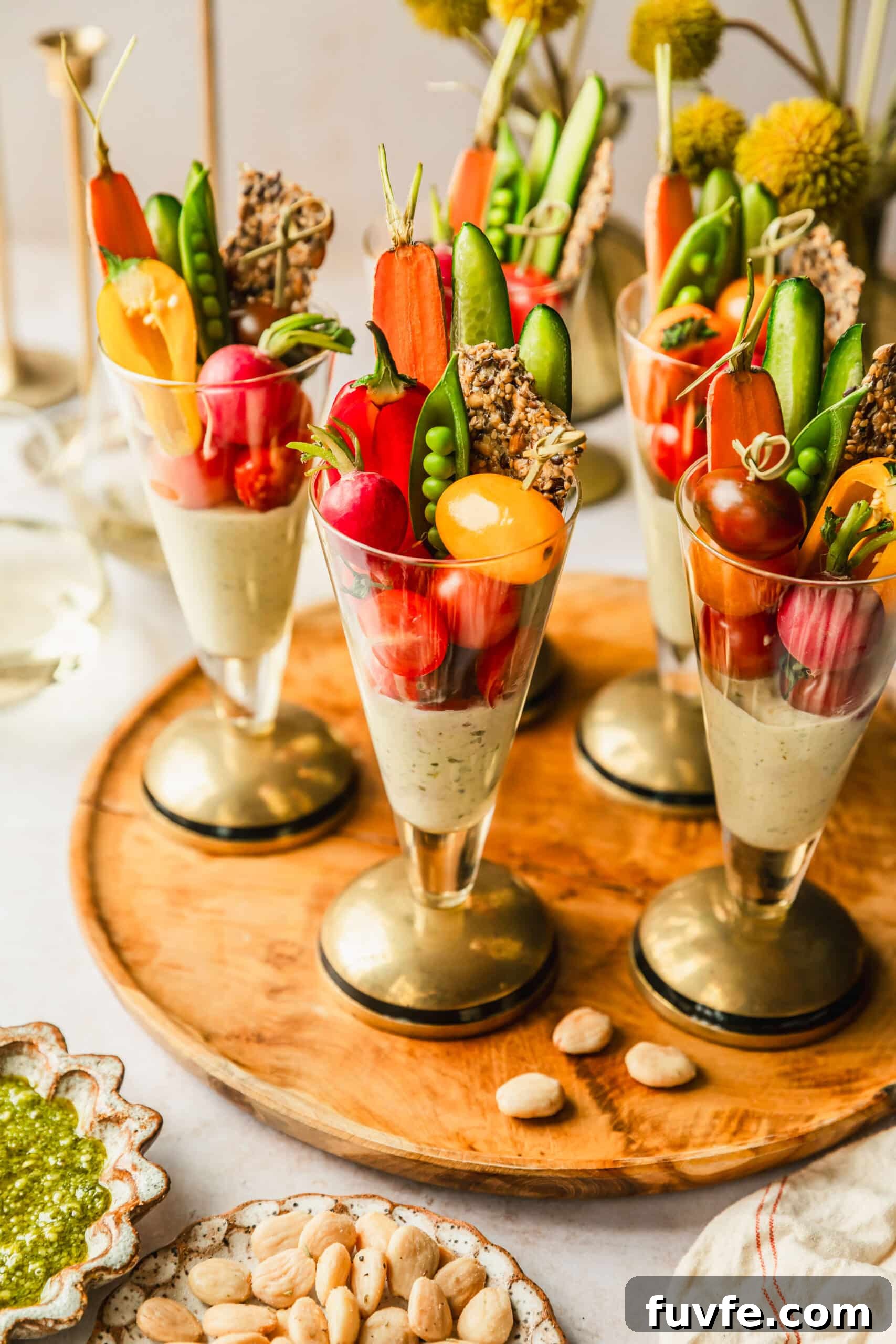 Crudité Cups with Fresh Pesto Dip for Entertaining 2 Crudités cups with pesto dip on a wood board next to white and brown bowls of almonds and pesto, glasses of white wine, and yellow flowers.