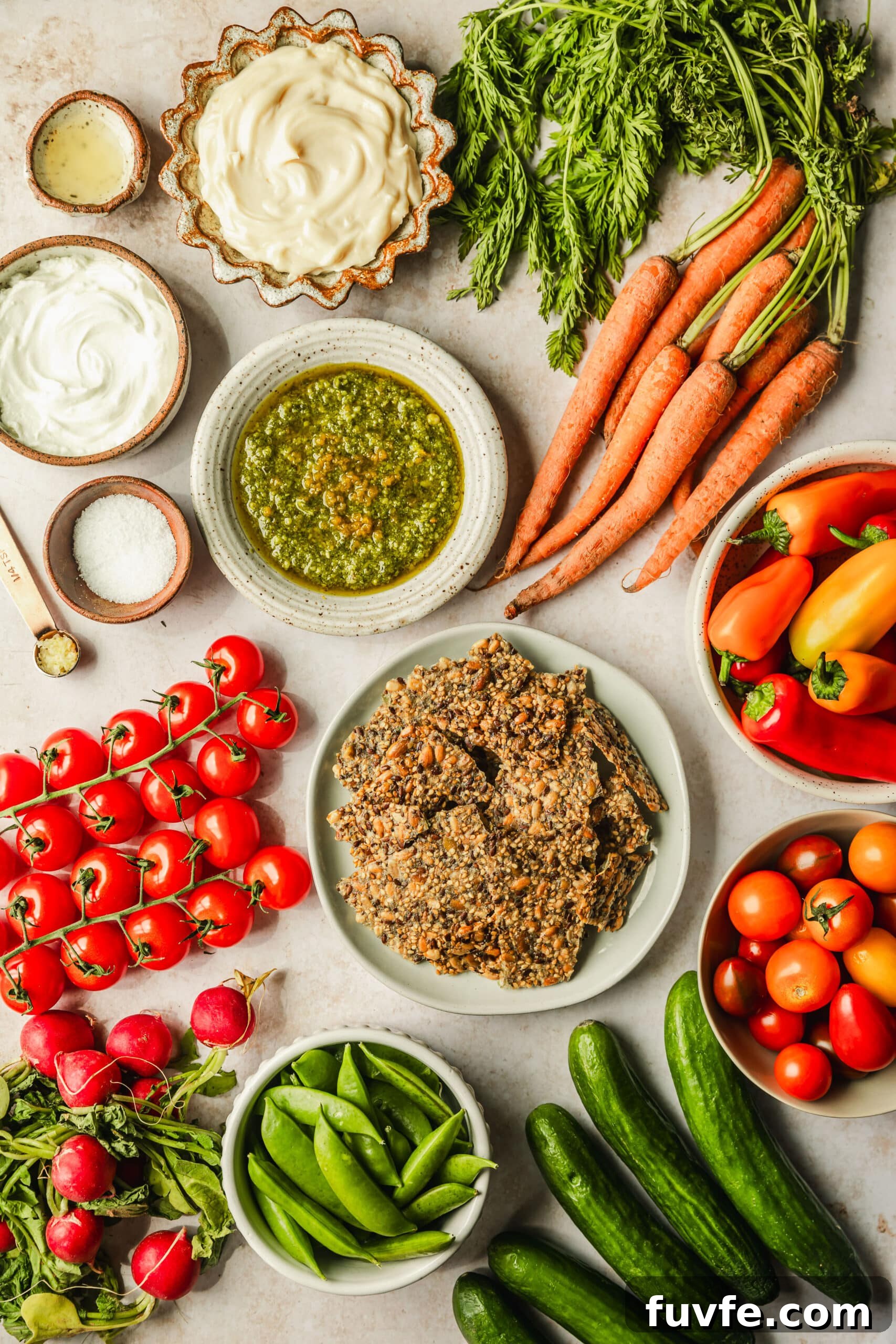 Crudité Cups with Fresh Pesto Dip for Entertaining 3 White and brown bowls of basil sauce, mayo, yogurt, salt, garlic, lemon juice, crackers, cherry tomatoes, peppers, and peas on a tan counter next to mini cucumbers, radishes, and carrots.