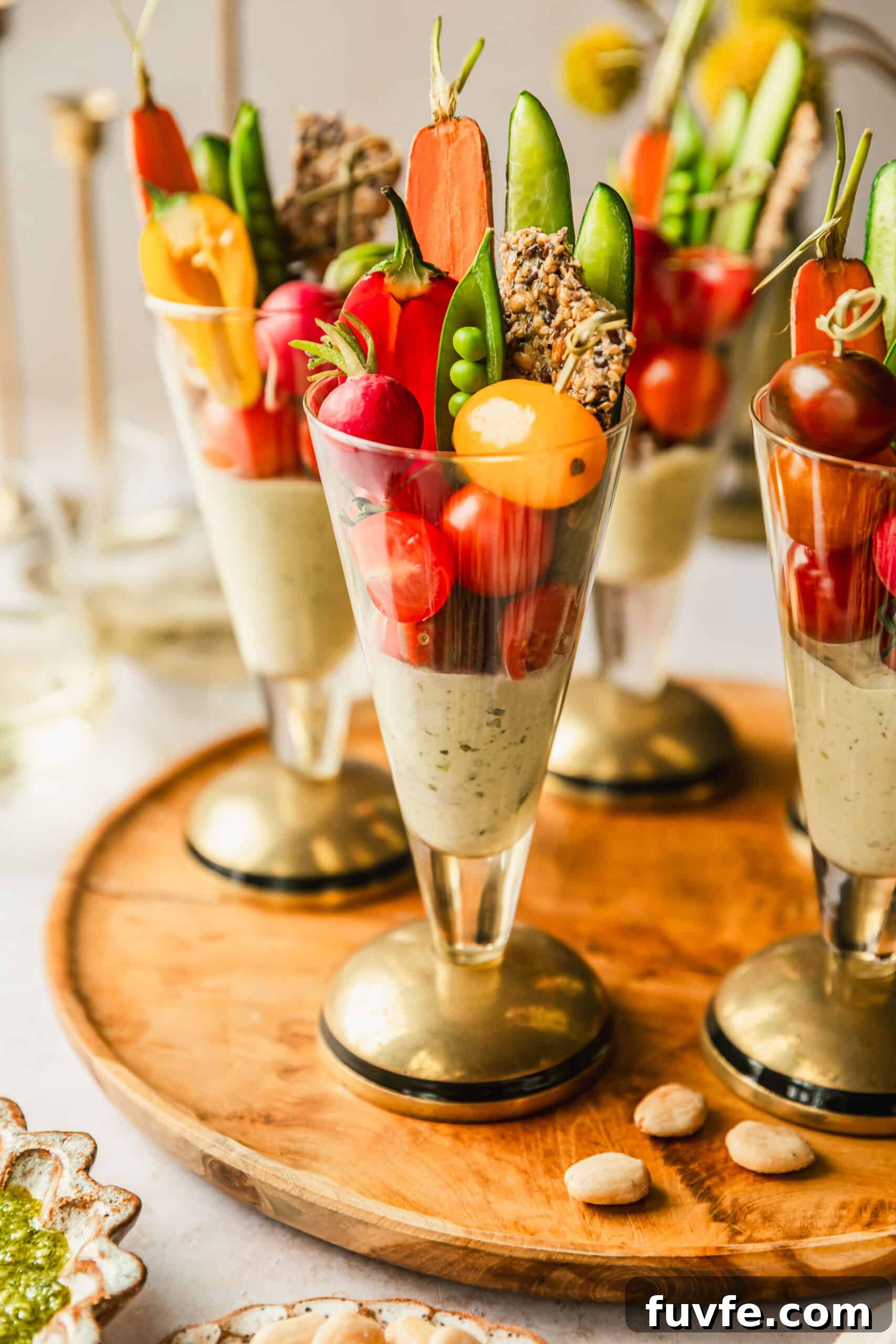 Crudité Cups with Fresh Pesto Dip for Entertaining 6 A wood tray of veggie cups with basil sauce on a tan counter next to glasses of white wine, a vase of yellow flowers, and a white plate of almonds.