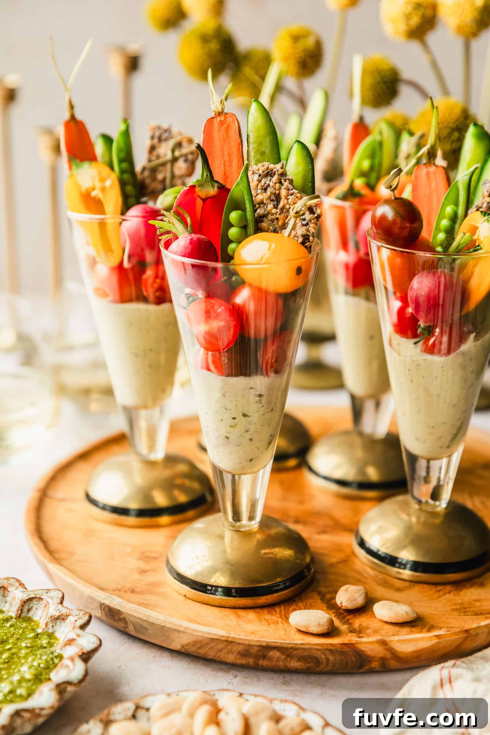Crudité Cups with Fresh Pesto Dip for Entertaining 7 A wood board of crudités cups with pesto dip on a tan counter next to yellow flowers, and white bowls of pesto and almonds.