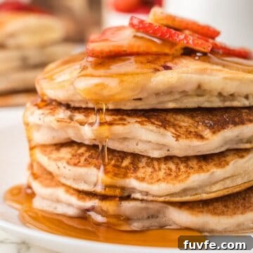 Four pancakes stacked on top of each other with syrup dripped down the sides and sliced strawberries on top.