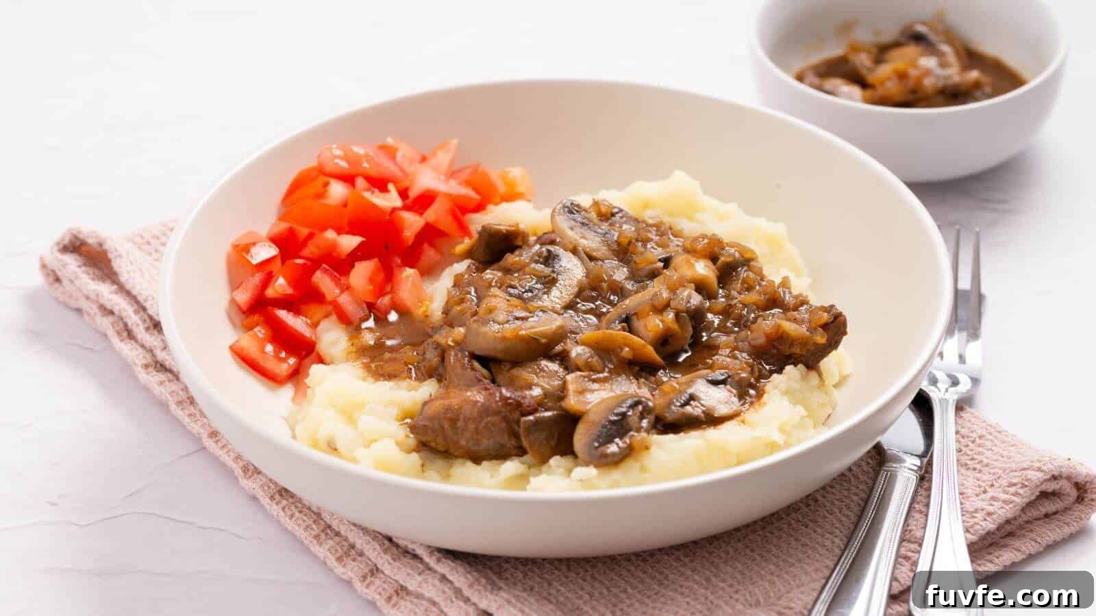 Plate with mashed potatoes topped with beef and mushroom gravy, alongside diced tomatoes. A bowl of extra gravy is in the background, with a fork and knife beside the plate.