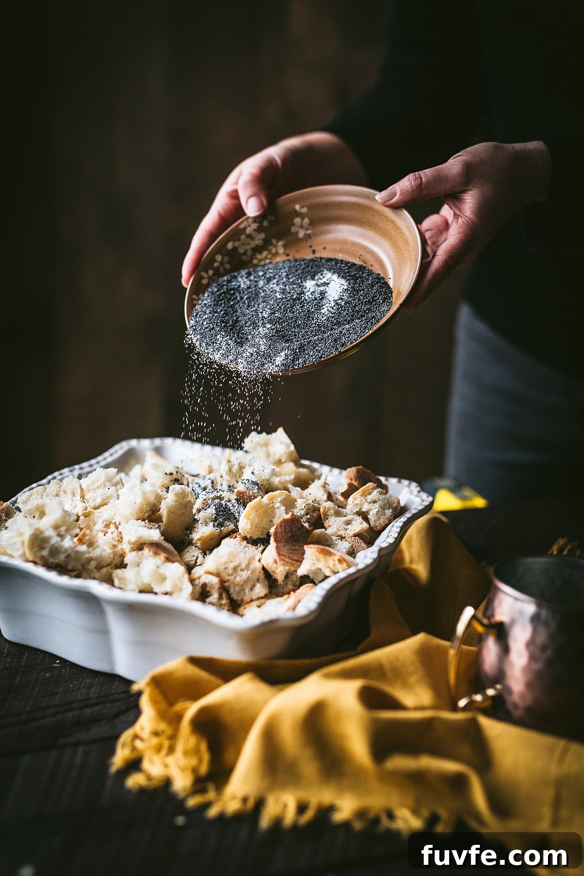 A woman pouring poppy seeds on top of a torn bread pieces in a white ceramic dish to make Mákos Guba