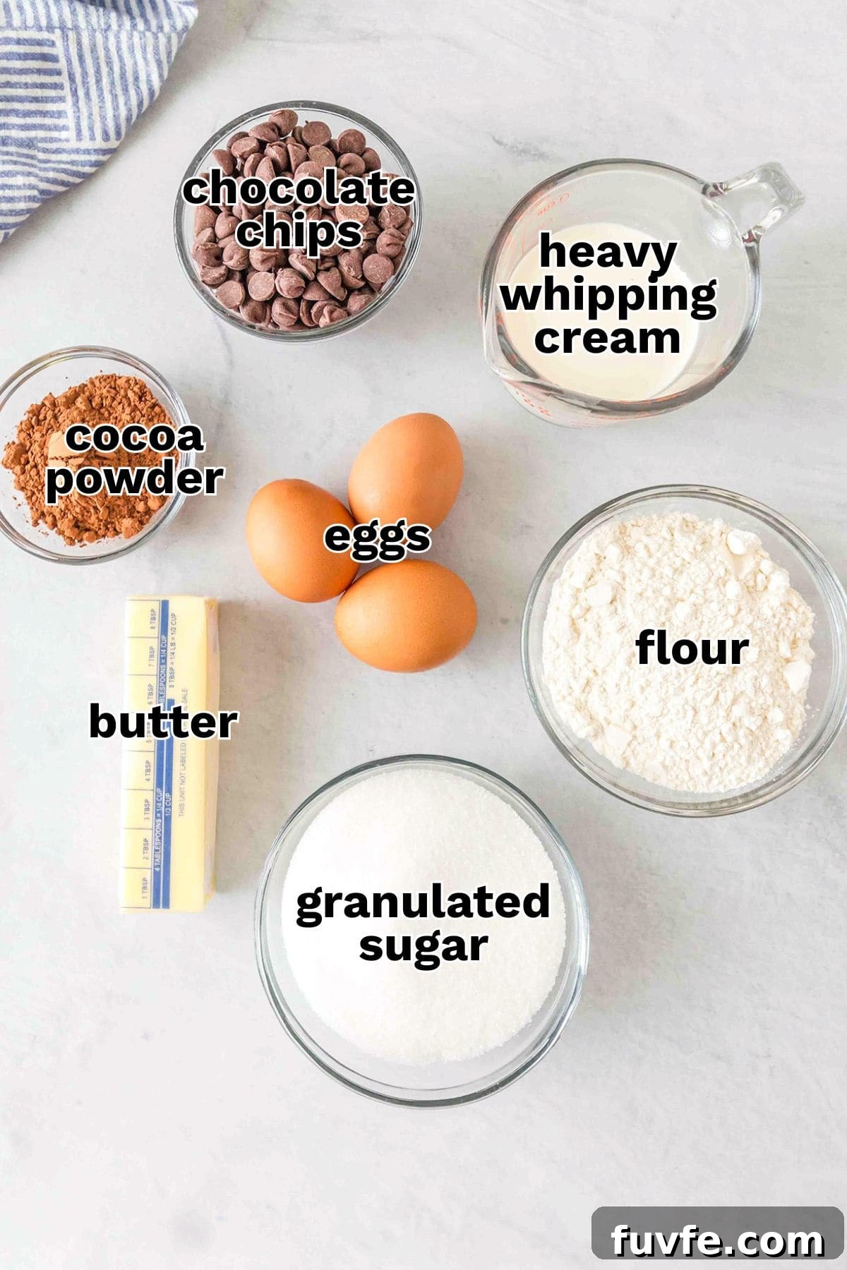 Decadent Chocolate Molten Lava Cake Recipe for Home Bakers 3 Six labeled ingredients for Chocolate Lava Cake—chocolate chips, heavy whipping cream, cocoa powder, eggs, butter, flour, and granulated sugar—are arranged on a white surface.