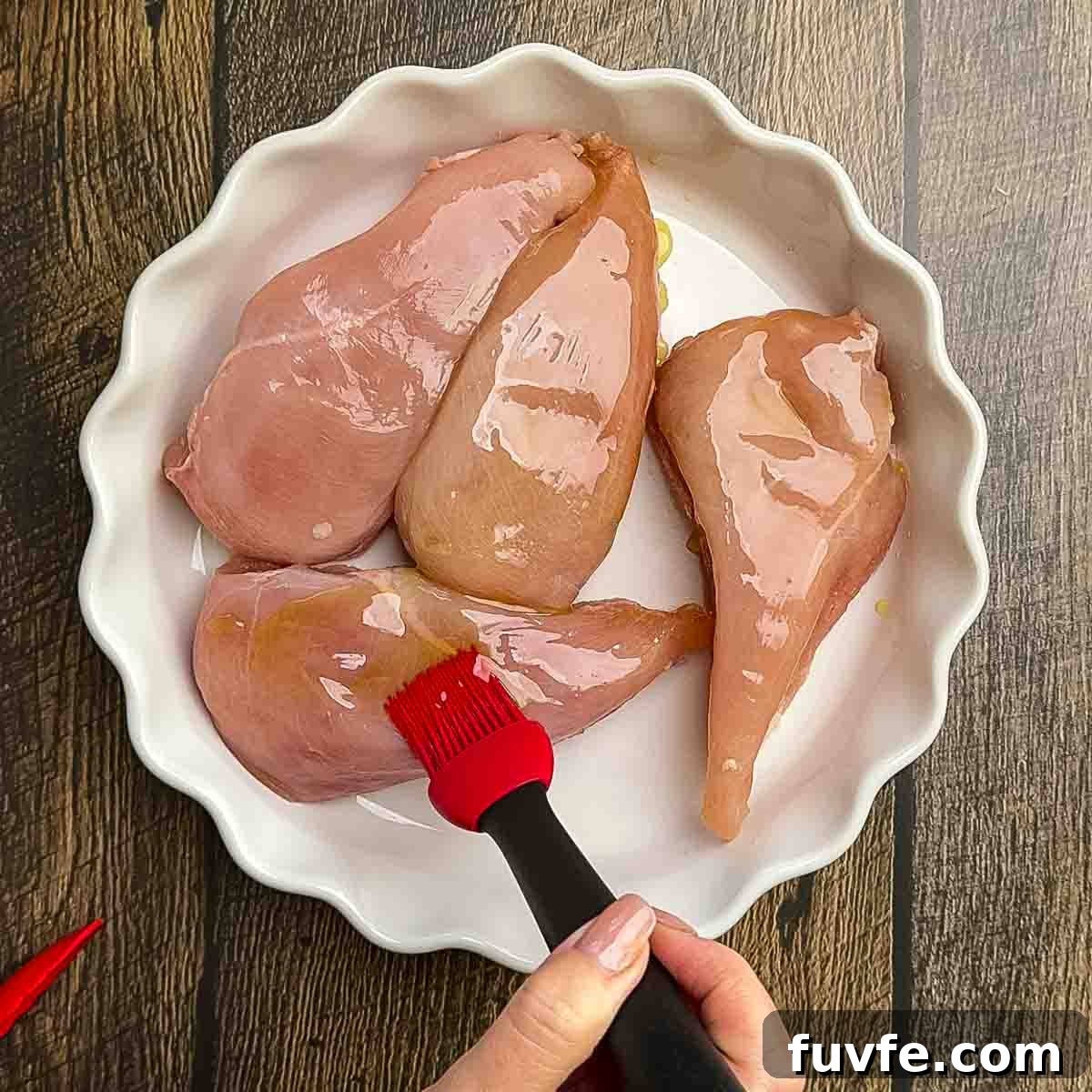 A red basting brush brushing olive oil on the boneless chicken breasts.