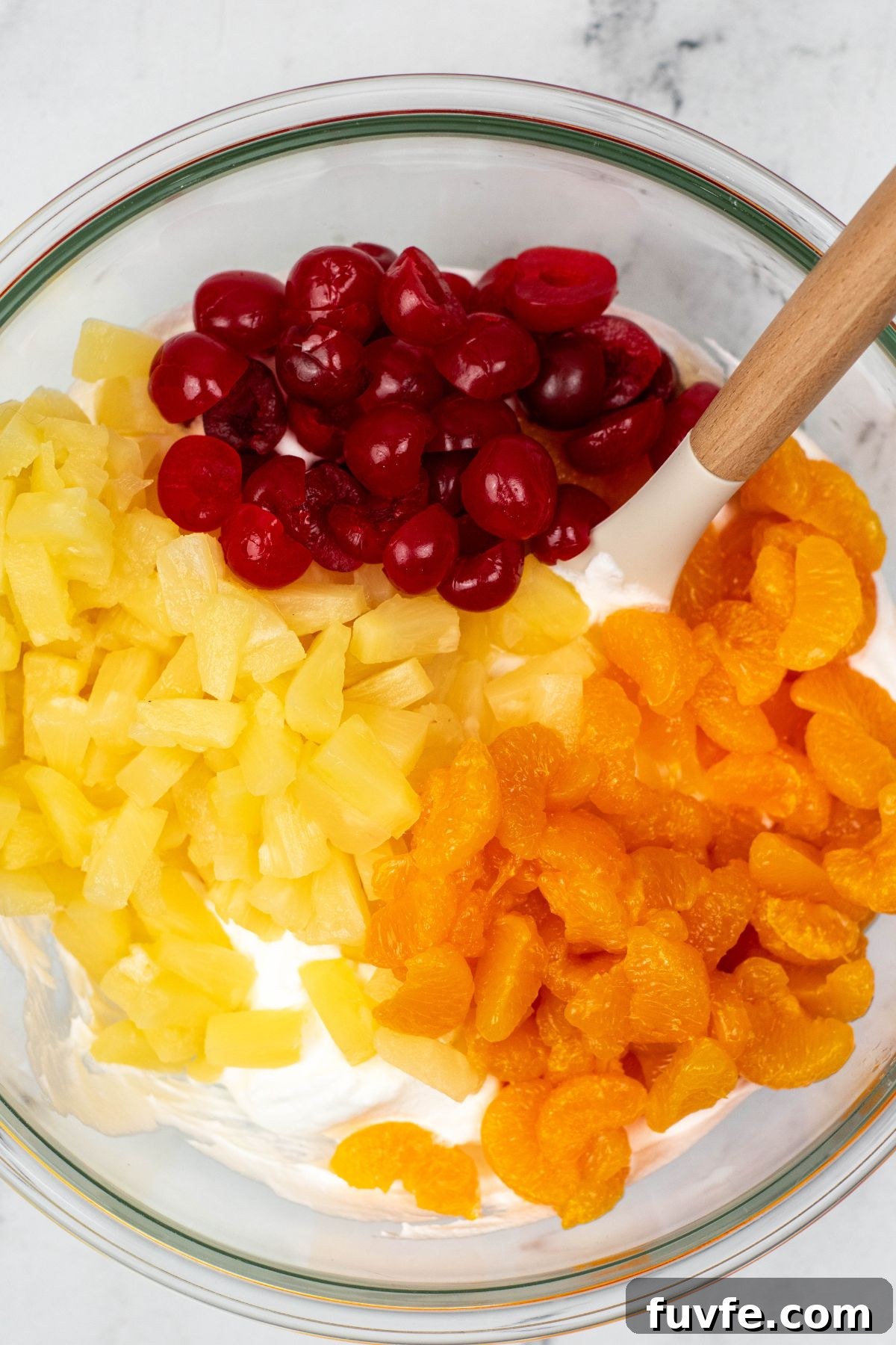 Cherries, pineapple, and mandarin oranges, being added to whipped cream mixture, in a clear glass bowl. 