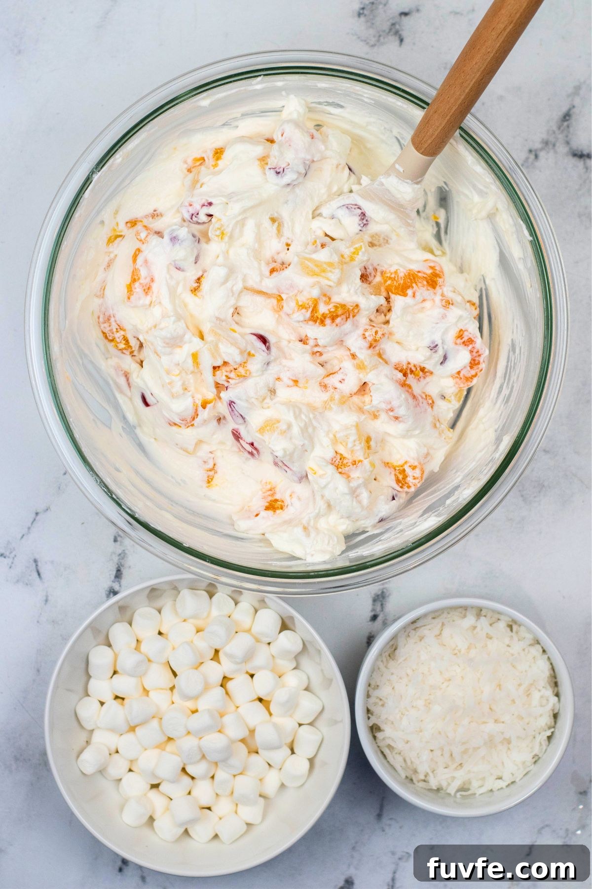 A large clear glass mixing bowl, filled with whipped cream mixture and fruit, having mini marshmallows and coconut. 