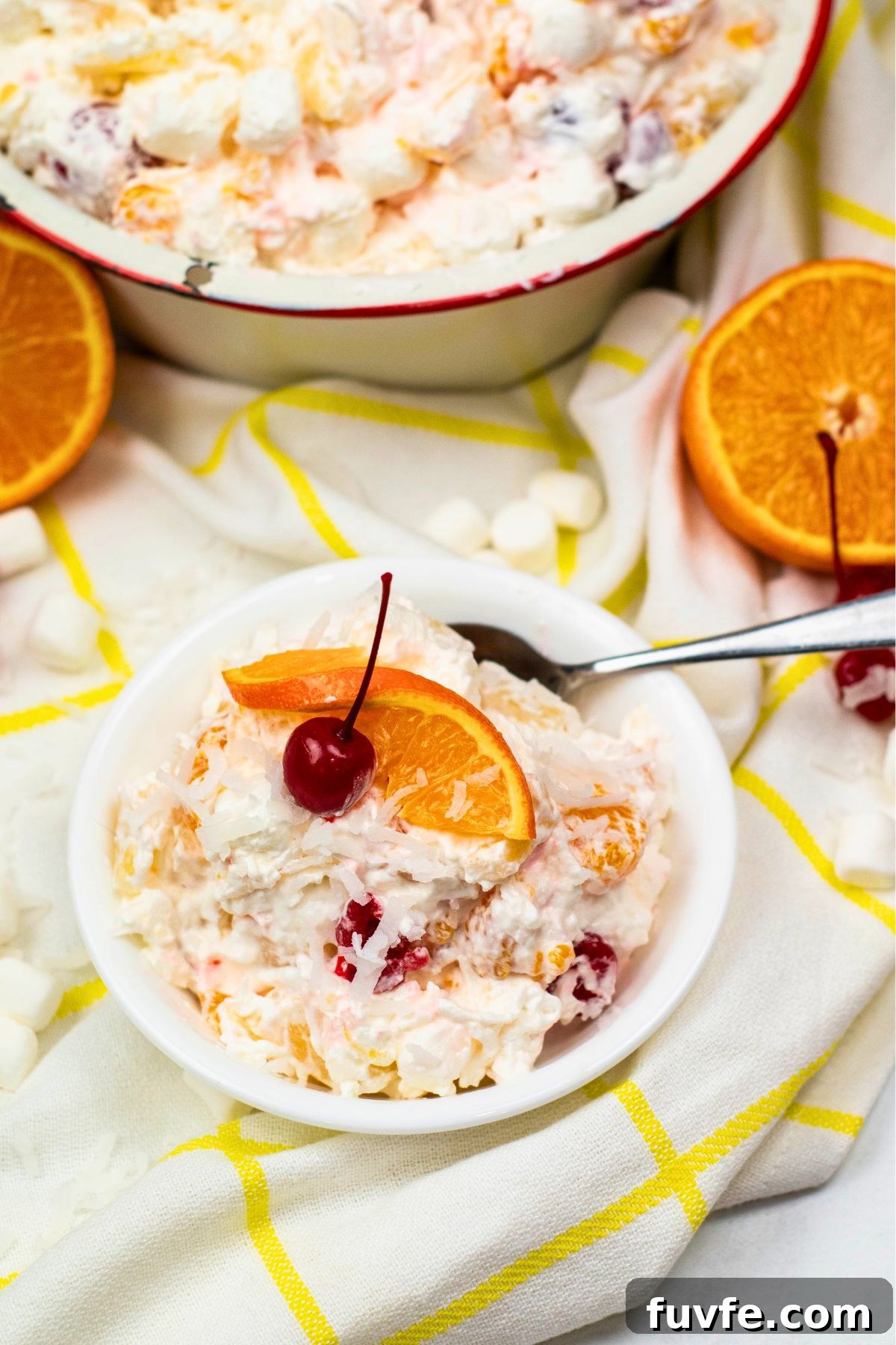 Small white bowl with ambrosia fruit salad in the bowl, along with a spoon. 
