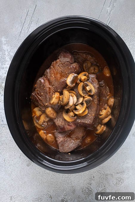 Slow cooker with browned beef, mushrooms and beef broth mixture.