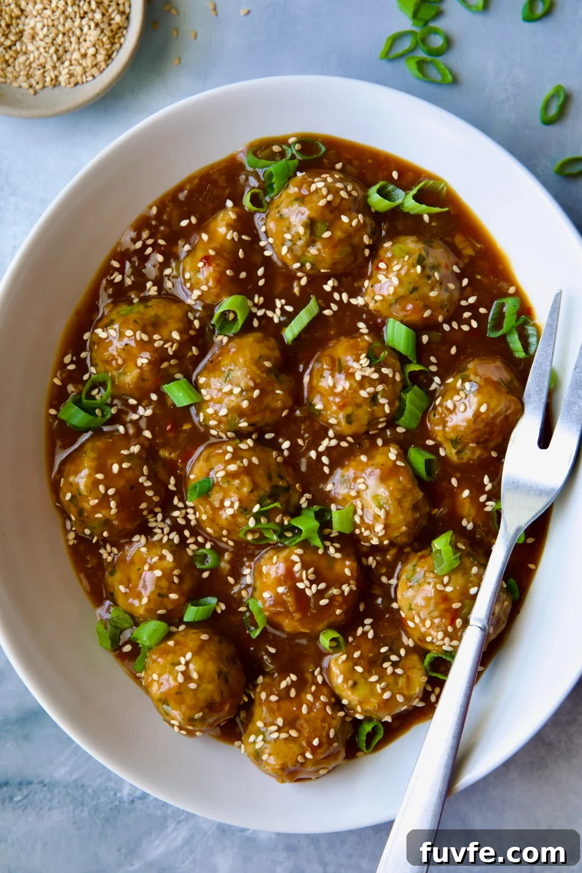 Asian-Style Turkey Meatballs with Ginger-Soy Glaze 2 Asian Turkey Meatballs in white oval serving bowl with serving fork garnished with sesame seeds and scallions.