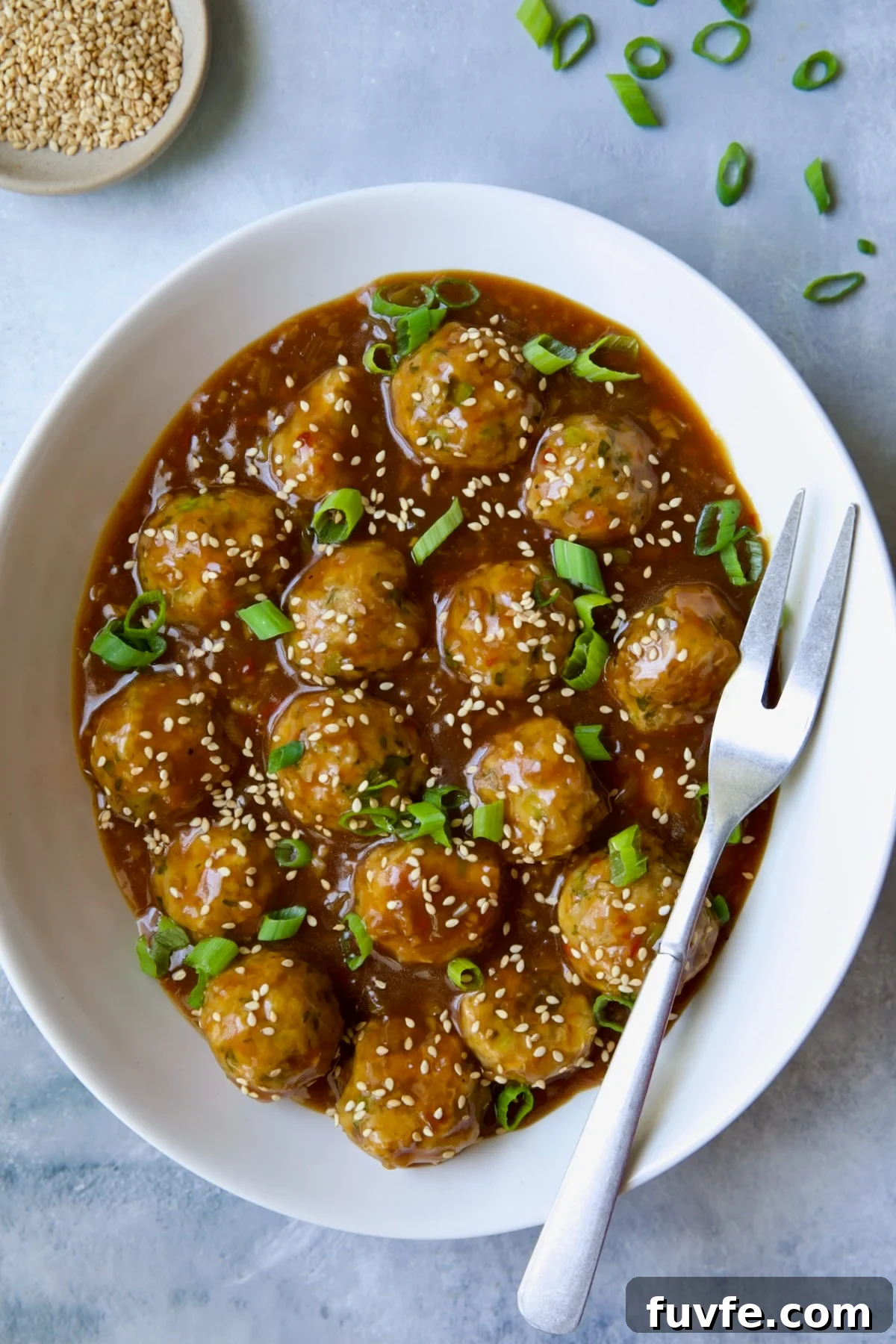 Asian-Style Turkey Meatballs with Ginger-Soy Glaze 13 Asian Turkey Meatballs in white oval serving bowl with serving fork garnished with sesame seeds and scallions.