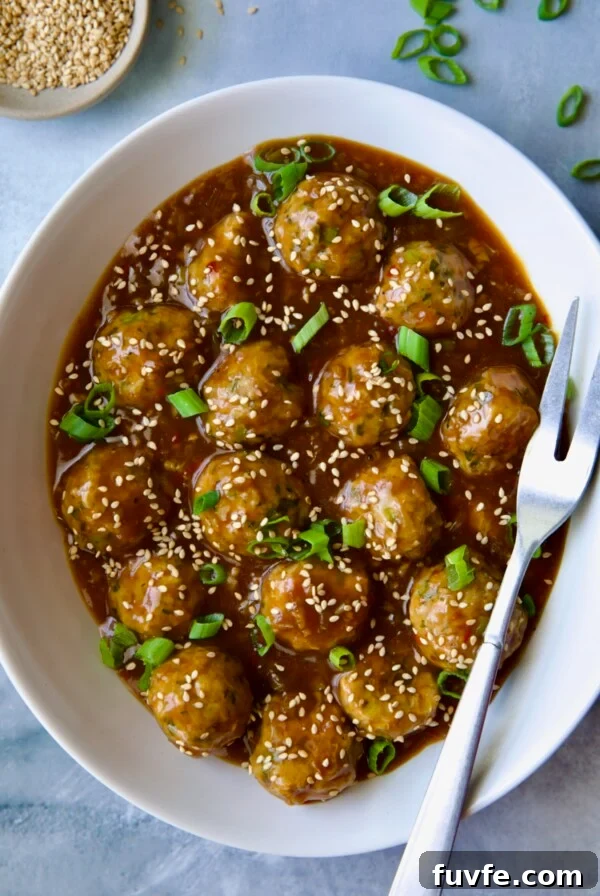 Asian-Style Turkey Meatballs with Ginger-Soy Glaze 16 Asian Turkey Meatballs in white oval serving bowl with serving fork garnished with sesame seeds and scallions.