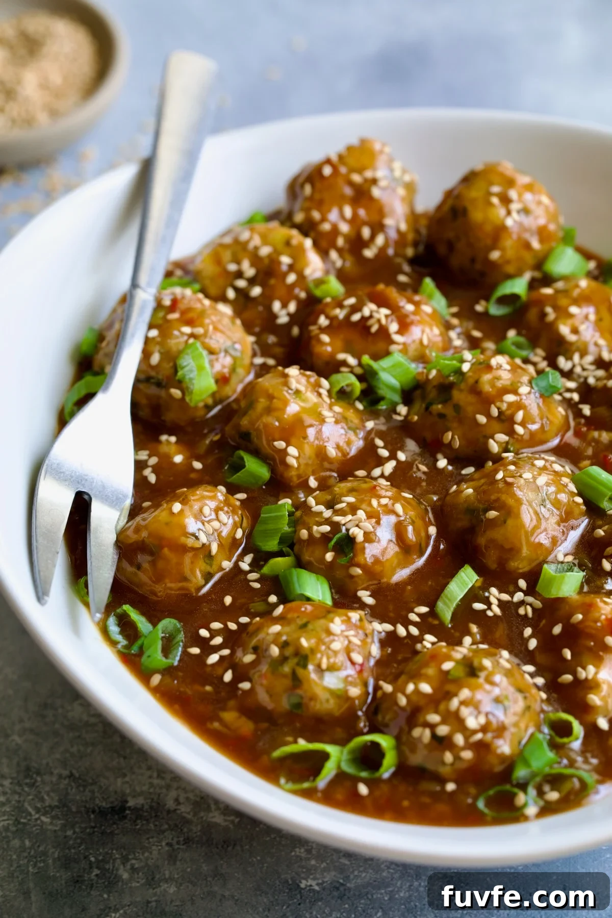 Asian-Style Turkey Meatballs with Ginger-Soy Glaze 3 Asian Turkey Meatballs in white oval serving bowl with serving fork garnished with sesame seeds and scallions.