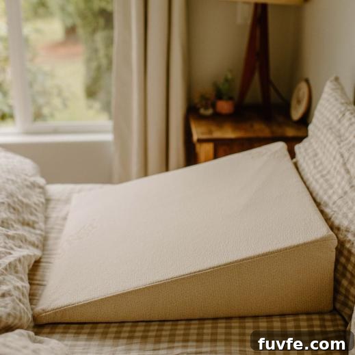 Top Non-Toxic Pregnancy and Organic Nursing Pillows for Moms 12 A large, cream-colored wedge pillow sits on a neutral-toned checkered bedsheet in a cozy room with a wooden bedside table and natural light coming through a window.