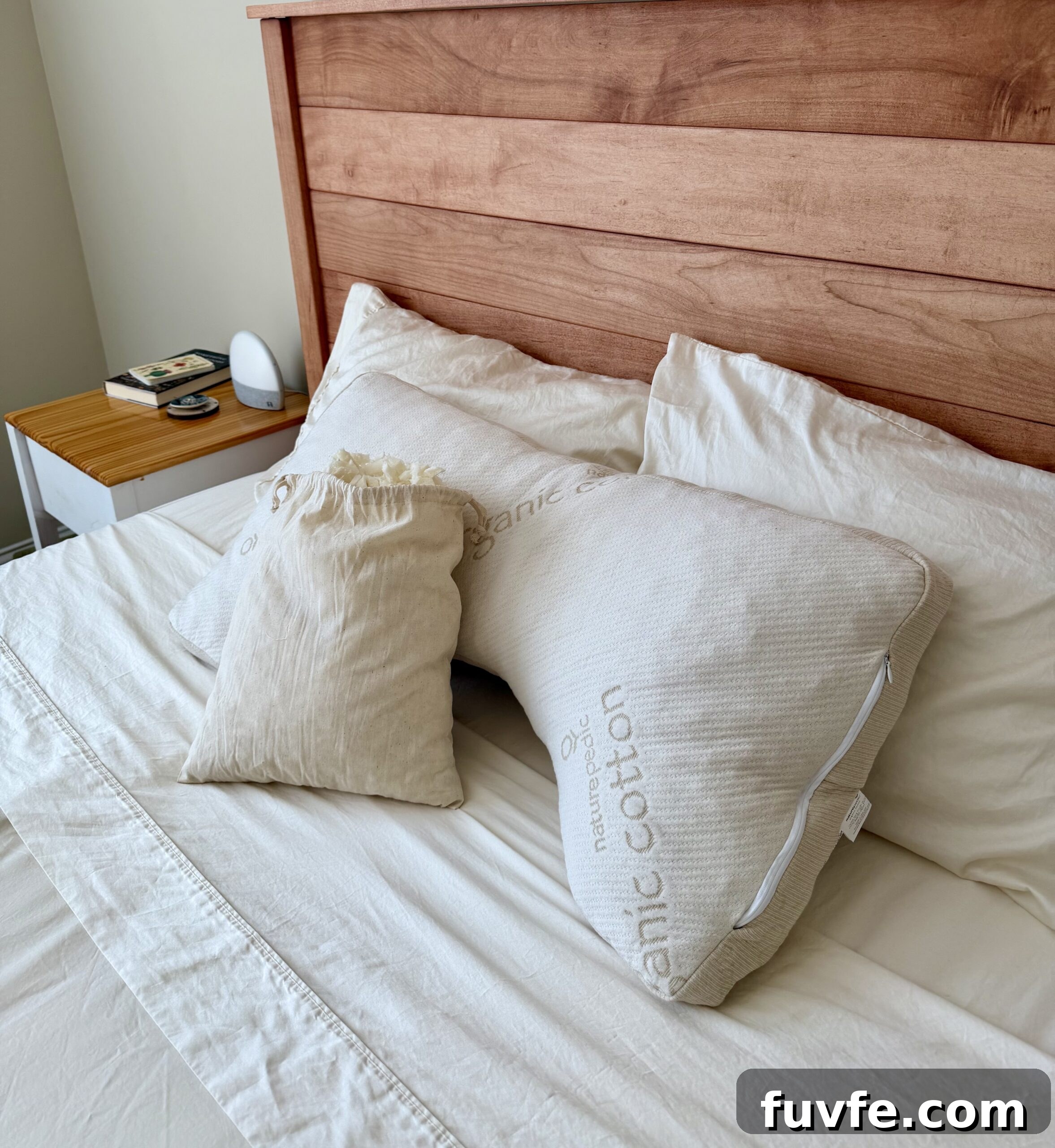 Top Non-Toxic Pregnancy and Organic Nursing Pillows for Moms 14 A neatly made bed with cream-colored sheets, two large pillows labeled organic cotton, and a smaller drawstring bag resting on top. A wooden headboard and a bedside table with books and a clock are visible.