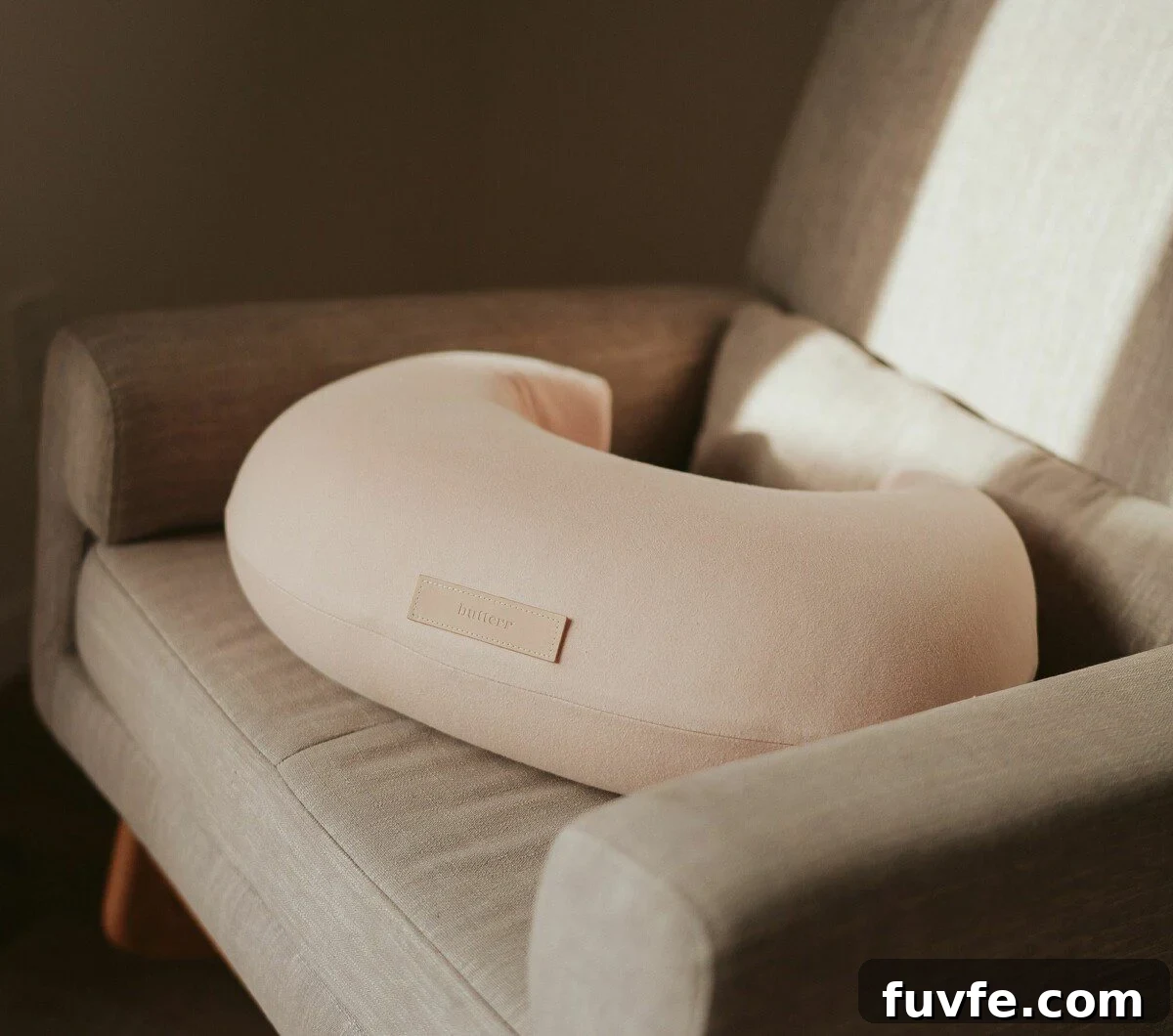 Top Non-Toxic Pregnancy and Organic Nursing Pillows for Moms 3 A peach-colored, crescent-shaped nursing pillow with a small leather brand tag rests on a neutral-toned armchair in a softly lit room.