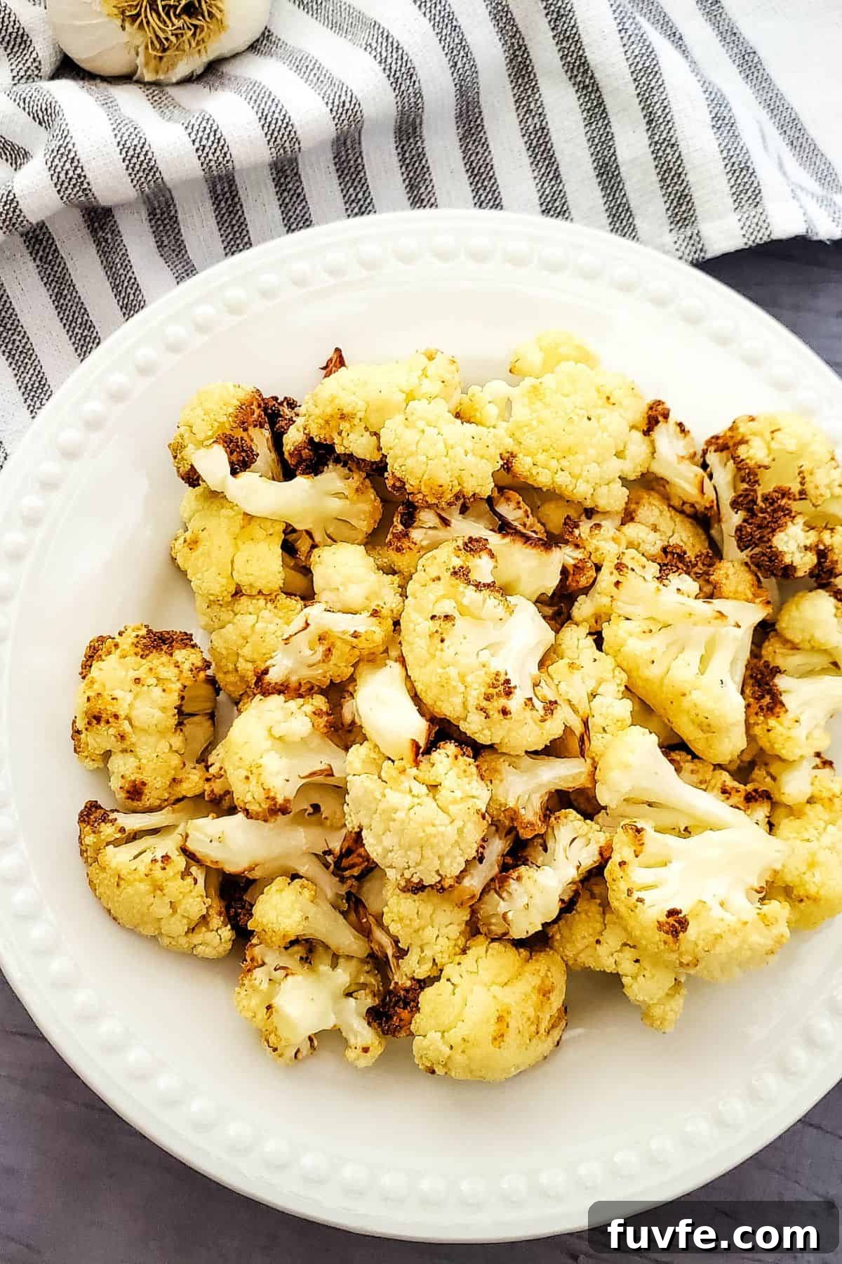 Crispy Air-Fried Cauliflower Recipes for Every Meal 3 A white plate with florets, set on a gray surface with a striped cloth and garlic visible in the background.