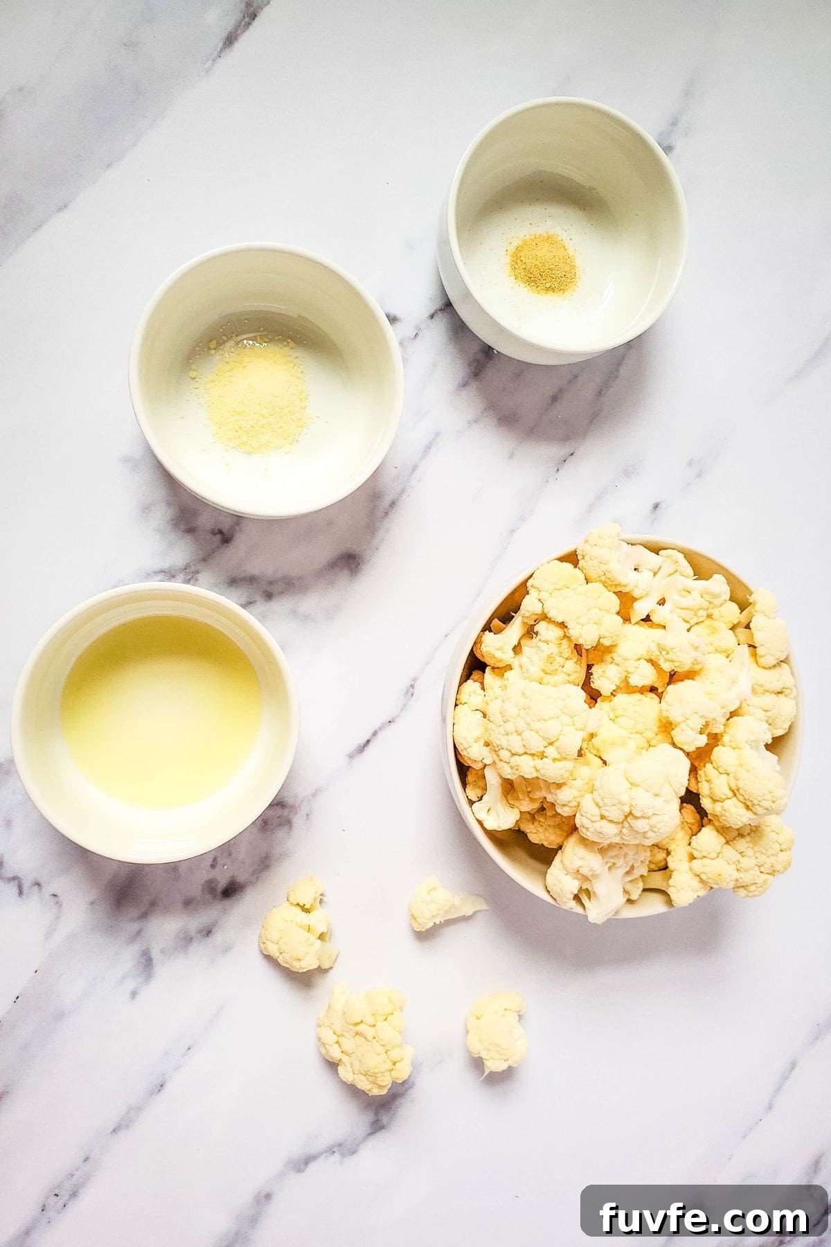 Crispy Air-Fried Cauliflower Recipes for Every Meal 4 Four bowls on a marble surface hold cauliflower florets, oil, grated cheese, onion powder, and garlic powder—ingredients perfect for making crispy air fryer fried cauliflower. A few scattered florets add to the inviting prep scene.