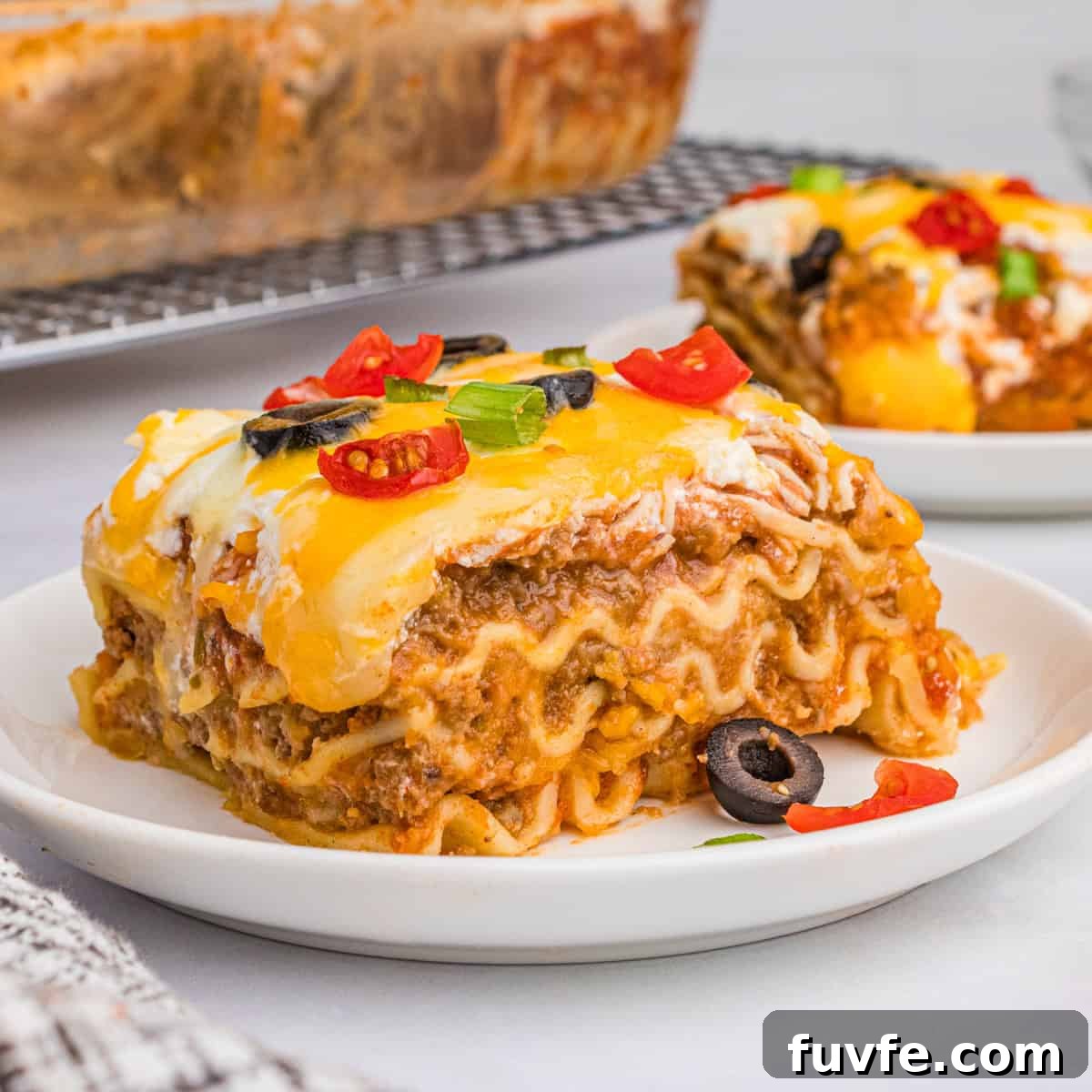 A slice of taco lasagna with noodles on a white plate, topped with melted cheese, olives, tomatoes, and green onions.