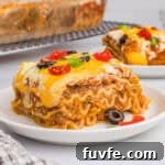 A slice of taco lasagna with noodles on a white plate, topped with melted cheese, olives, tomatoes, and green onions.