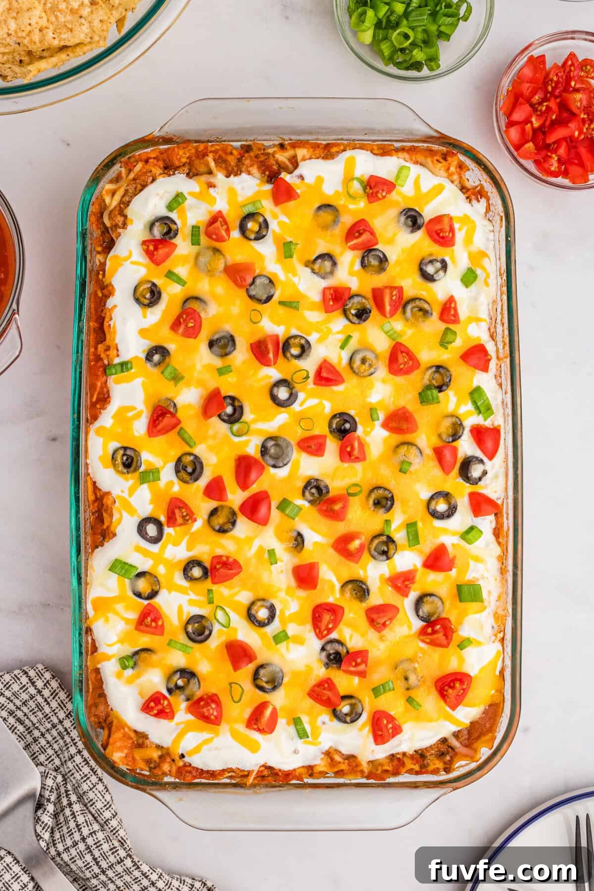 Taco lasagna in a glass 9 x 13 baking dish topped with sour cream, cheese, olives, tomatoes, and green onions.