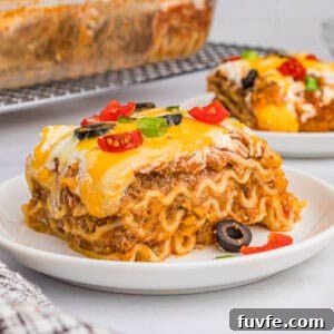 A slice of taco lasagna with noodles on a white plate, topped with melted cheese, olives, tomatoes, and green onions.