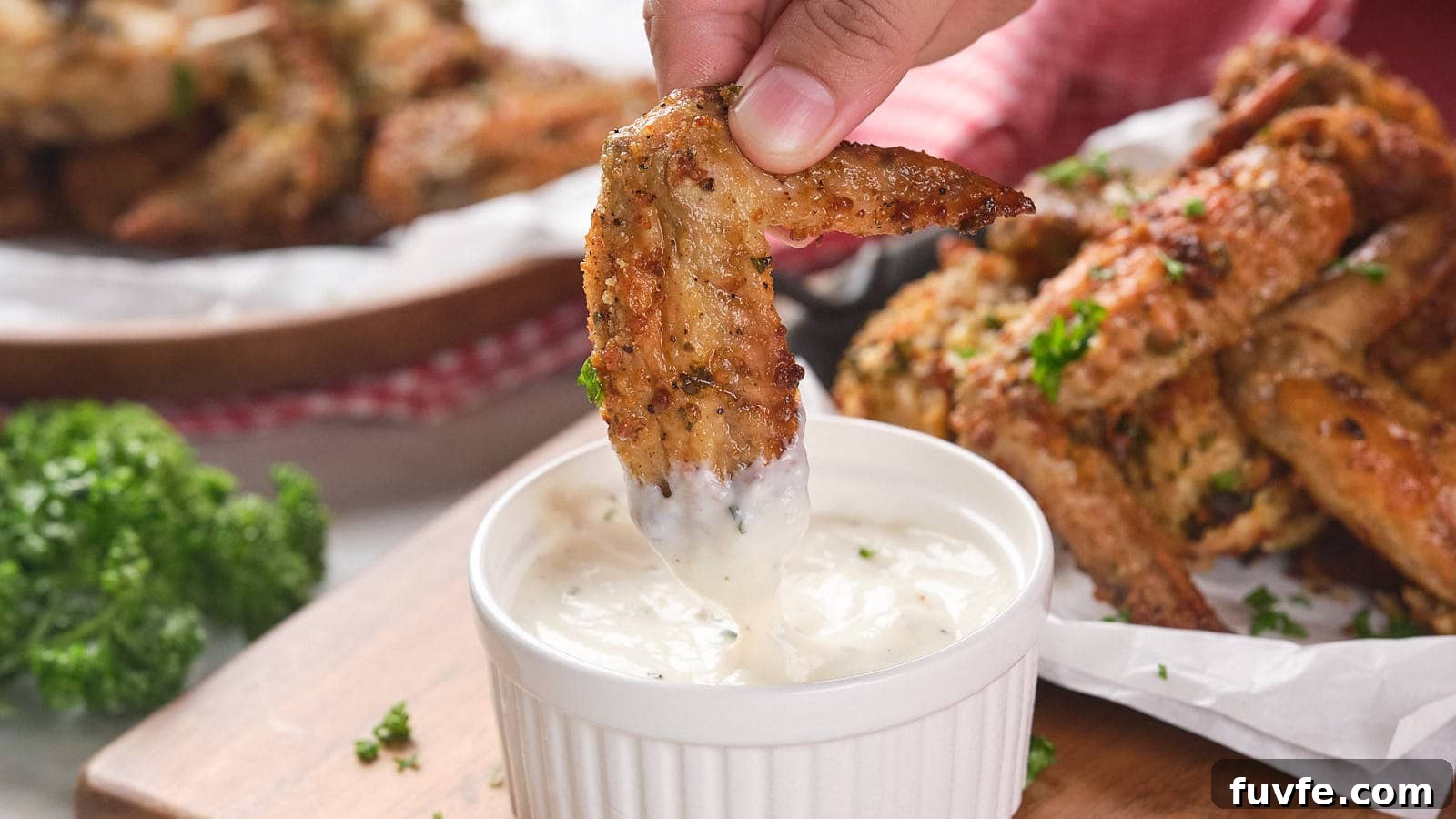 A hand dips a parmesan chicken wing into a small ramekin filled with white dipping sauce.
