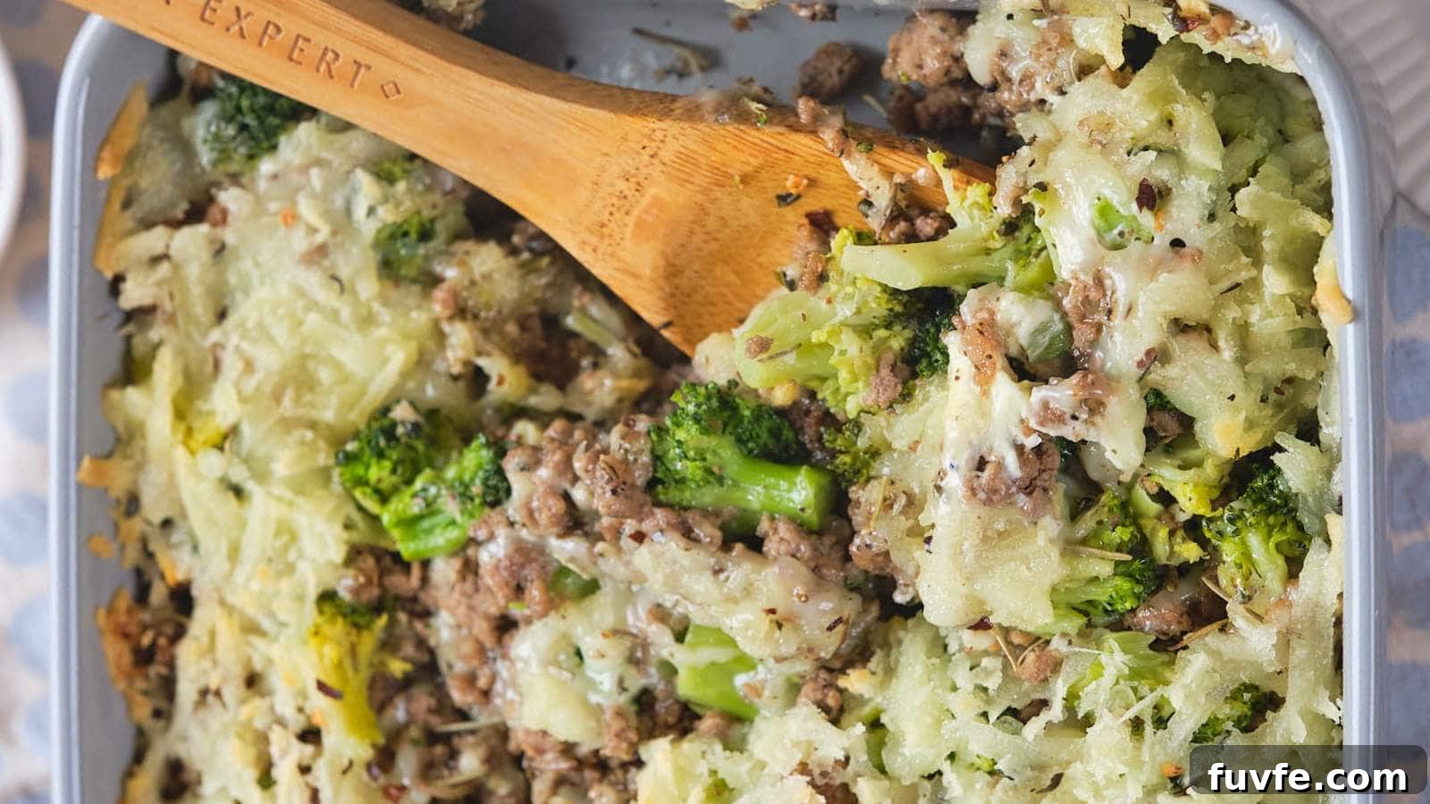 A wooden spoon rests in a dish of baked casserole containing ground meat, broccoli, and a shredded potato topping.