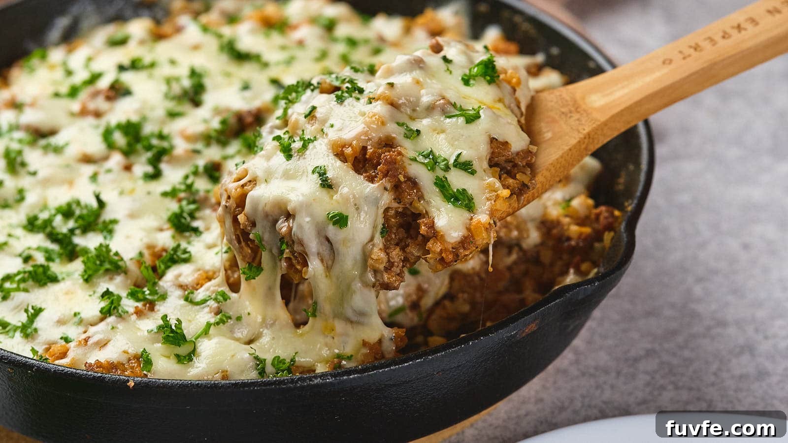 A wooden spoon lifting a serving of italian ground beef skillet garnished with parsley from a cast-iron skillet.
