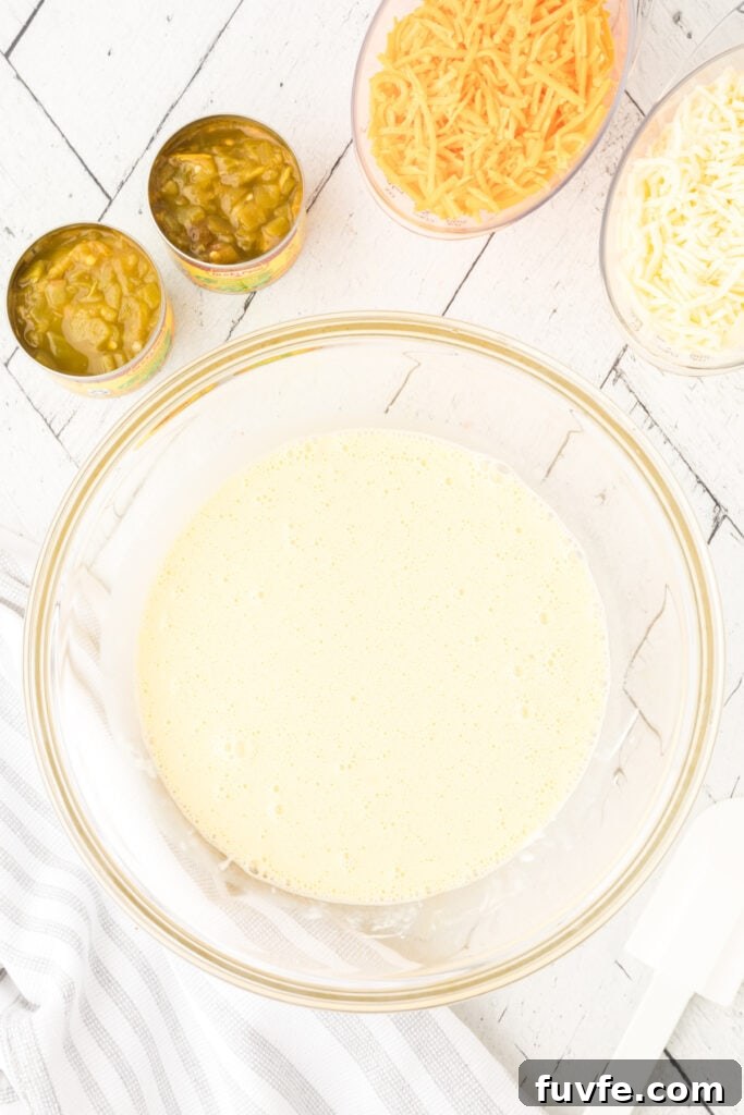 Whisked egg mixture in a glass bowl with green chilies and shredded cheese nearby