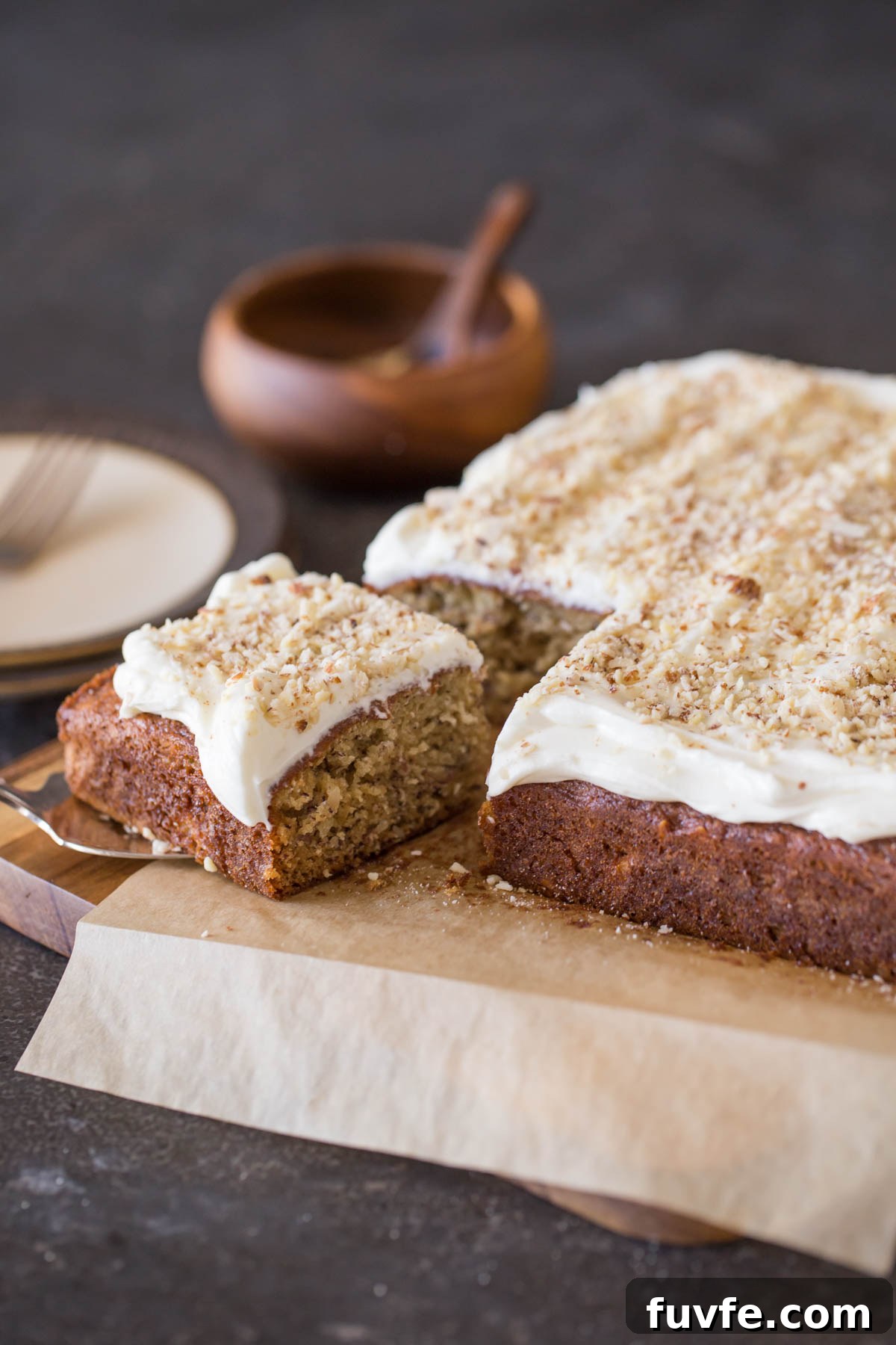 Banana Cake with Fluffy Cream Cheese Frosting on parchment paper on top of a cutting board, with the corner piece on top of a serving spatula. 